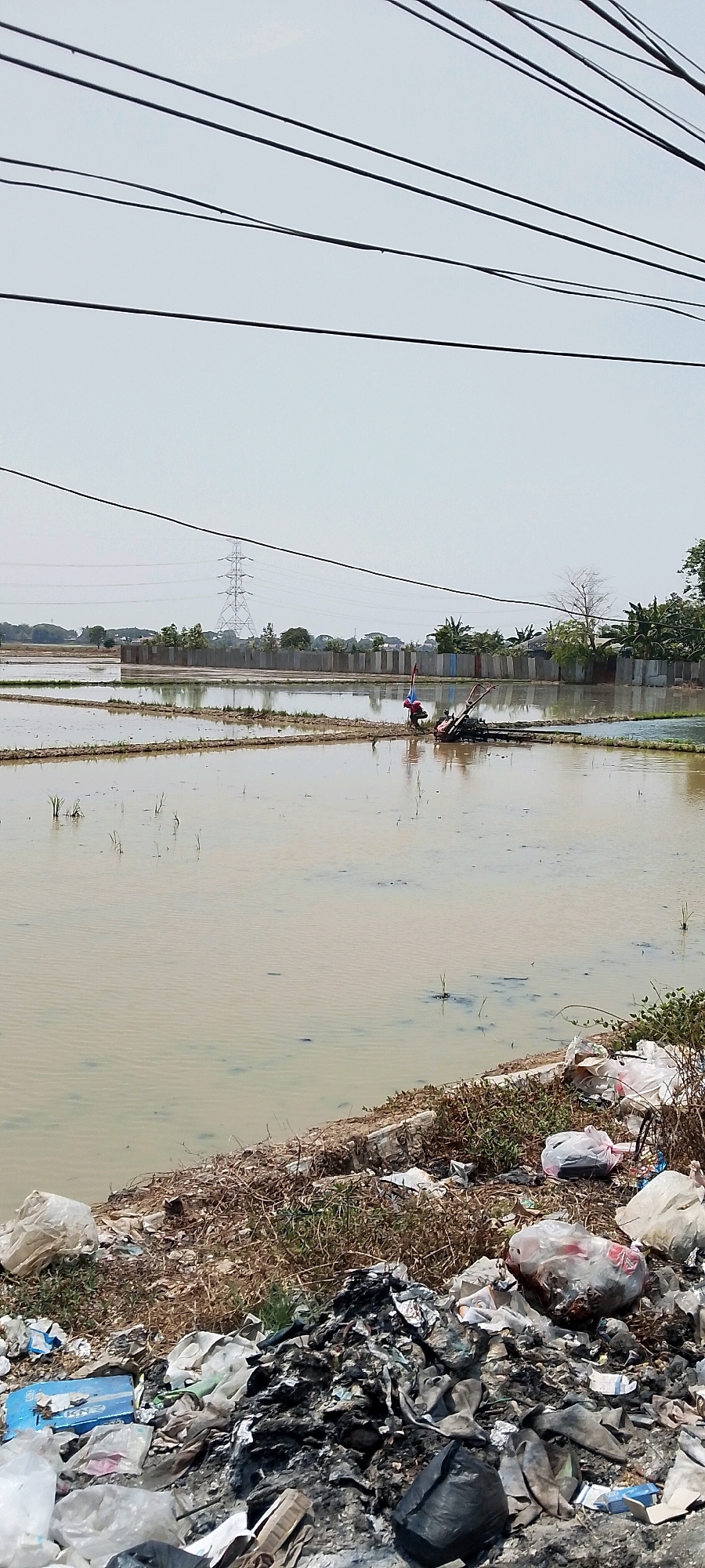 In Sukaraja, Indonesia, a rural scene unfolds with a flooded rice paddy occupying the majority of the frame. The murky water reflects the overcast sky, and sparse green shoots of rice are beginning to emerge. In the mid-ground, a person, possibly a farmer, is operating a small, red-topped tiller in the water. Beyond the paddy, a utilitarian concrete barrier stretches horizontally, with a transmission tower visible in the distance. The foreground is marred by a significant accumulation of refuse, including plastic bags, debris, and what appears to be burnt material, creating a stark contrast with the natural elements. High-tension wires are strung across the top of the image, dividing the sky.
