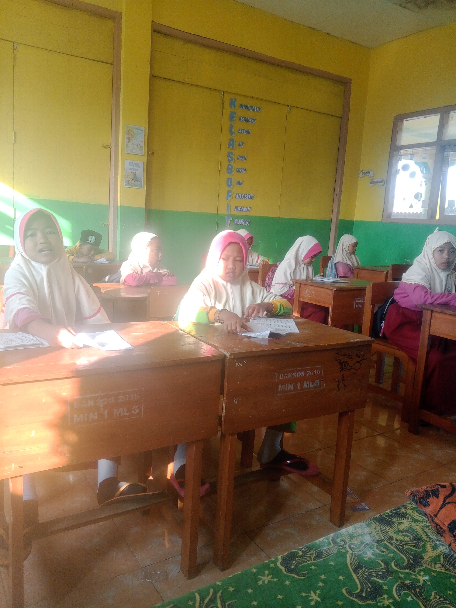 In a brightly lit classroom in Kemulan, Indonesia, several young girls wearing headscarves are seated at wooden desks, engaged in learning activities. Sunlight streams in from the left, casting long shadows across the room and illuminating the yellow walls. The desks have various markings, including "BAKSOS 2018" and "MIN 1 MLG," suggesting they are part of a school. One girl in the foreground on the left is looking directly at the camera with a slightly open mouth, holding a piece of paper. To her right, another girl is focused on a paper on her desk. Further back, several other girls are also seated at their desks, some looking forward and others downwards. In the background, a word cloud of Indonesian words like "KOMUNIKATIF," "KSPESIF," and "ESTARI" is displayed on the wall. A window is visible on the right side of the image, showing a blurry outdoor scene. The floor is tiled.