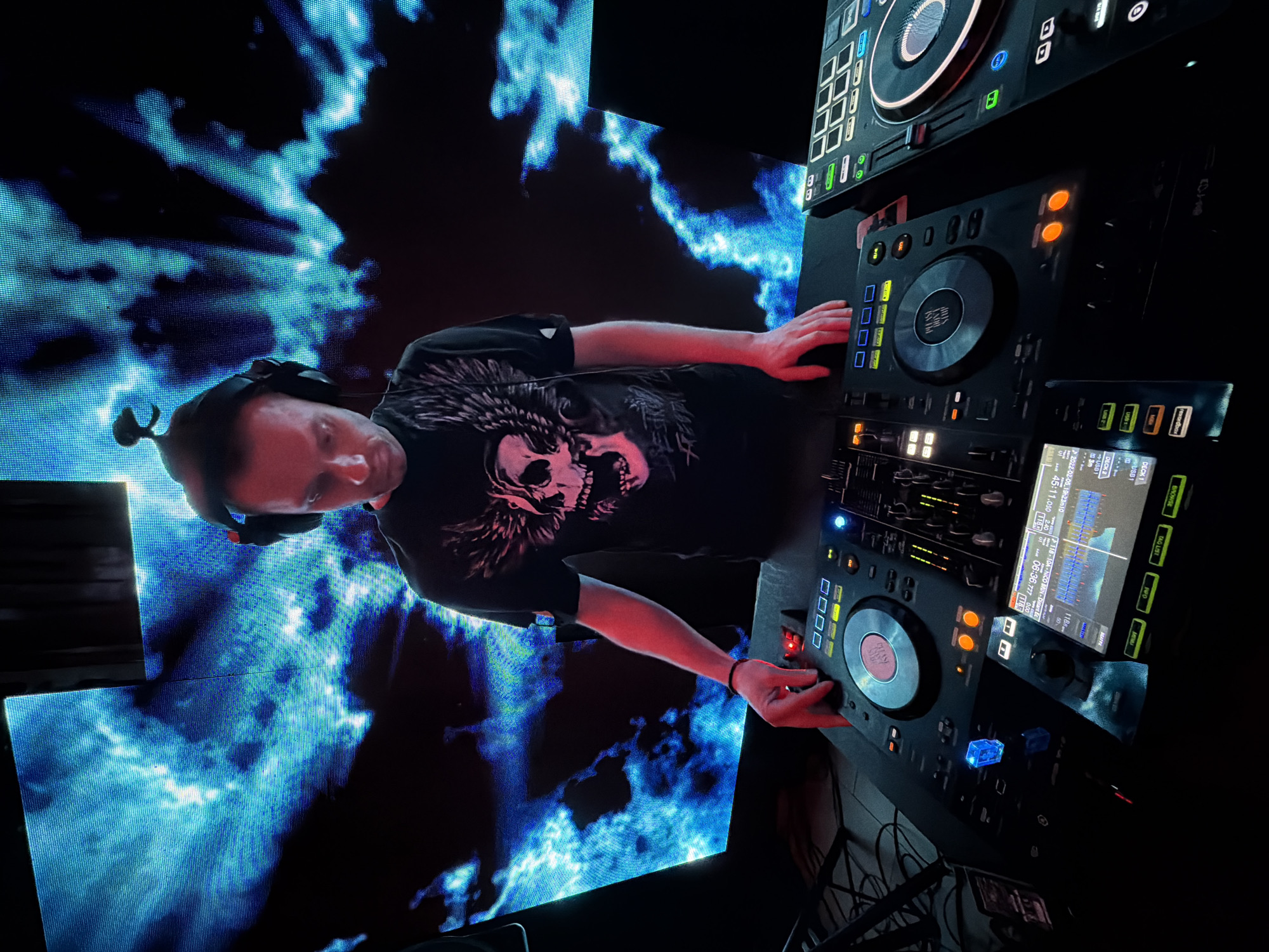 A man wearing headphones and a black t-shirt with a skull graphic is operating a DJ setup in what appears to be a nightclub or concert venue. He is facing the DJ equipment with his hands on the controls. Behind him, a large screen displays a blue and white swirling pattern. The DJ equipment includes turntables, mixers, and a display screen. The setting is dark, with most of the lighting coming from the screens and the DJ equipment's controls. It is impossible to tell if the picture was taken in Votkinsk, Russia.