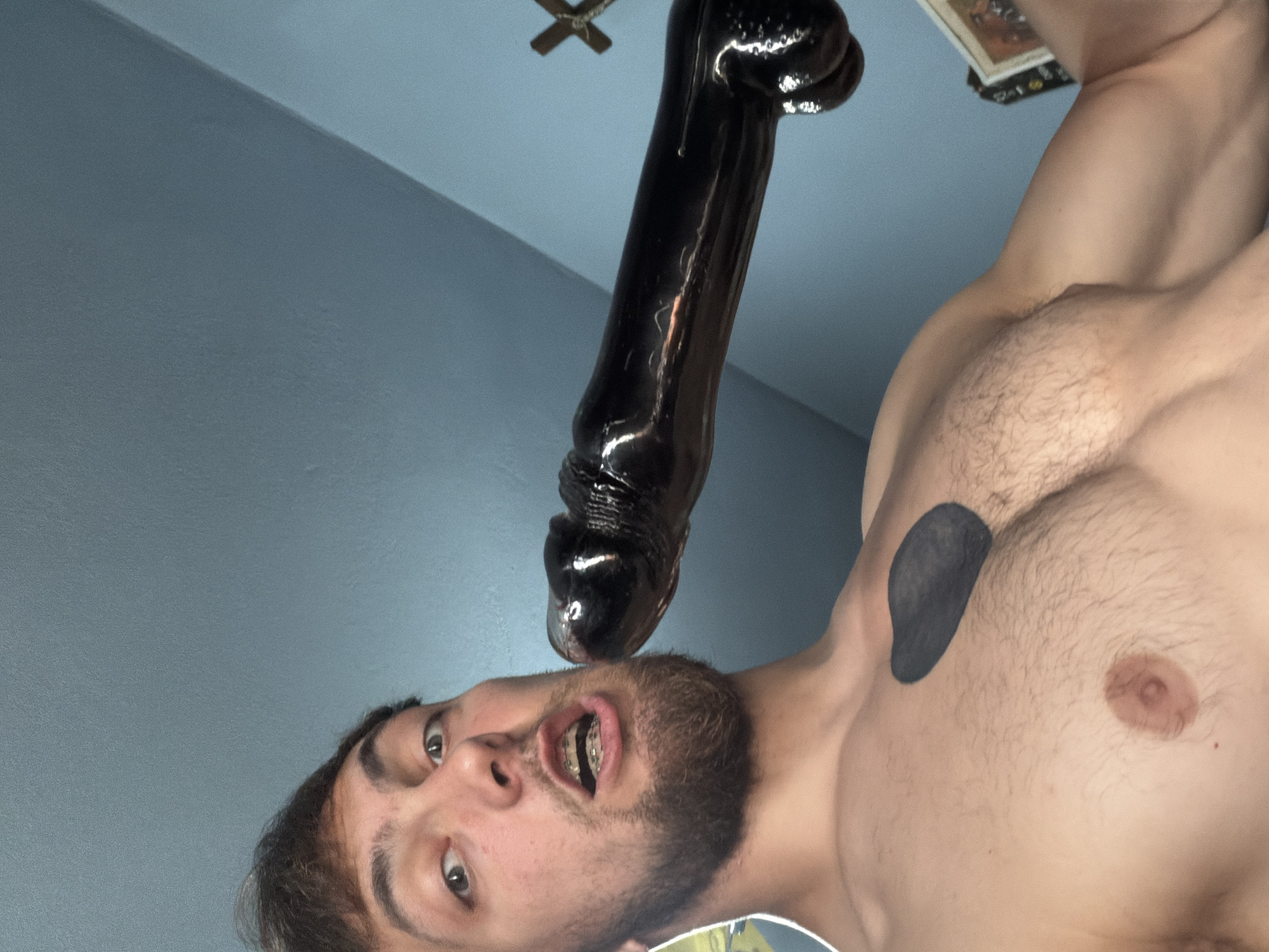 An upward-angled, close-up shot captures a shirtless man interacting with a large, dark, shiny object indoors, in what is contextually described as Tsarevo, Bulgaria.
The central figure is a young to middle-aged man with light skin, dark hair, and a short beard. He has braces on his upper teeth. His eyes are wide open, looking directly towards the viewer or camera, and his mouth is agape. A dark, irregularly shaped tattoo is visible on his left pectoral muscle. His chest and shoulders are muscular, with some body hair present.
Above his face, a prominent, long, and thick dark object is positioned, appearing to be held near or entering his open mouth. The object has a glossy, almost wet appearance, reflecting light in several places. Its texture seems somewhat irregular.
The background consists of a plain light blue wall or ceiling, suggesting an indoor setting. In the upper left corner, a partial view of a small ceiling fan is faintly discernible. In the upper right background, a book or similar small item is partially visible, but any text on it is unreadable. The overall lighting is soft and even. No specific time of day or weather conditions can be determined from the image. The man's expression is somewhat neutral, focused, or slightly surprised. No other visible text is present in the image.