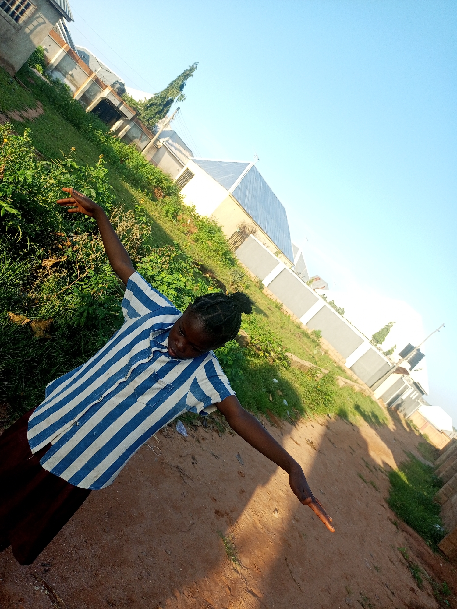 A young Black person with braided hair, wearing a blue and white striped short-sleeved shirt and a dark skirt, is standing with arms outstretched in a dirt path. They are looking down with a slight smile. The background features green grass, bushes, and buildings with grey and white walls under a clear blue sky. The image is slightly tilted. The location is identified as Tempure, Nigeria.