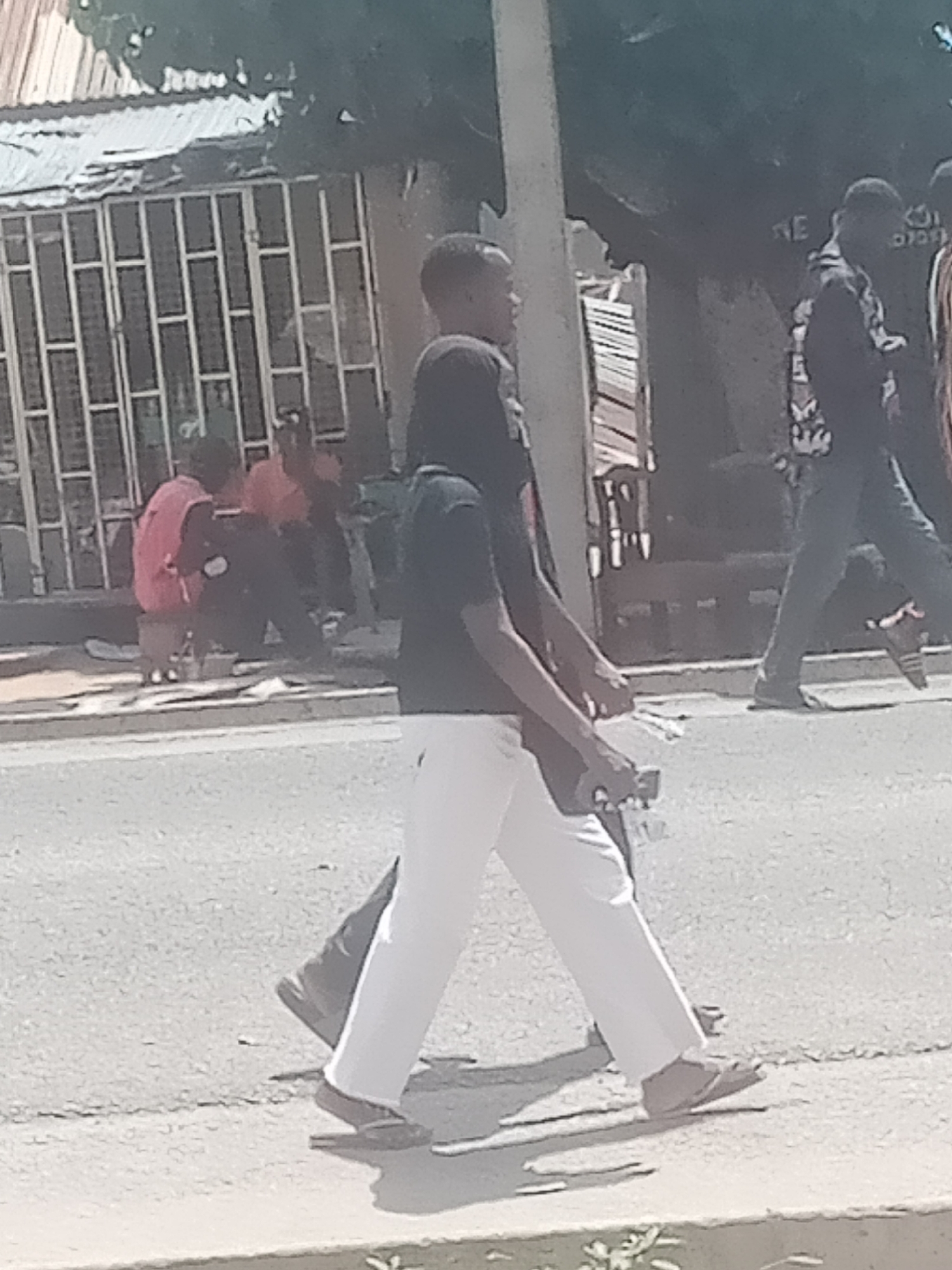 The image depicts a street scene in Jalingo, Nigeria. In the foreground, two individuals are walking on a paved road. The person on the left is wearing white trousers and dark footwear, and the person on the right is wearing dark trousers and dark footwear. Behind them, other people are visible in the background, some sitting and some standing, near buildings with corrugated metal roofs and barred windows. The environment appears to be an urban or semi-urban setting.