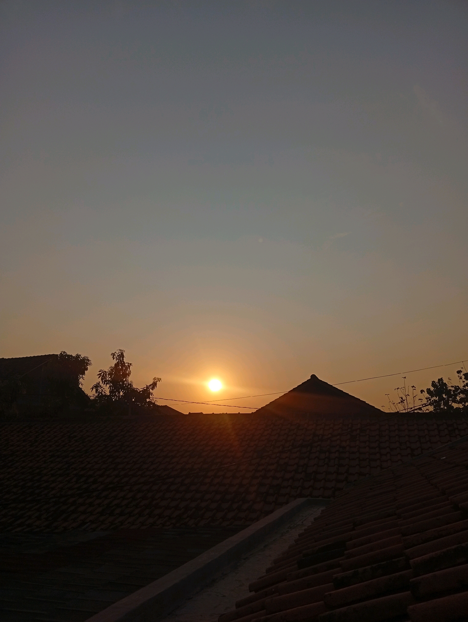 This is a vertical, low-angle shot of a sunset over a residential area in Kebandingan, Indonesia. The sky transitions from a pale blue at the top to a warm orange near the horizon. The sun, a bright, glowing orb, is partially obscured by trees and rooftops, casting a warm light across the scene. In the foreground, several tiled rooftops are visible, some in shadow and some catching the last rays of sunlight. A sharp, triangular roof peak is prominent in the mid-ground, with a thin power line extending across the sky. Silhouetted trees with sparse foliage are scattered throughout the image, adding depth and texture. The overall impression is one of a peaceful, twilight moment in an Indonesian neighborhood.