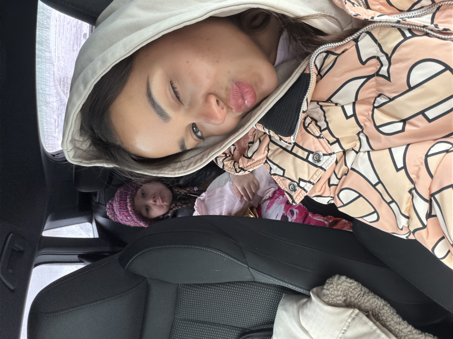 An angled close-up shows a woman wearing a hooded jacket, pursing her lips and winking, with a child wearing a purple knit hat visible behind her, seated in what appears to be a car seat. The car's interior, including a black leather seat, is also visible. The woman's jacket has a pattern of abstract, geometric shapes in shades of peach, beige, and black. The child seems to be looking directly at the camera.