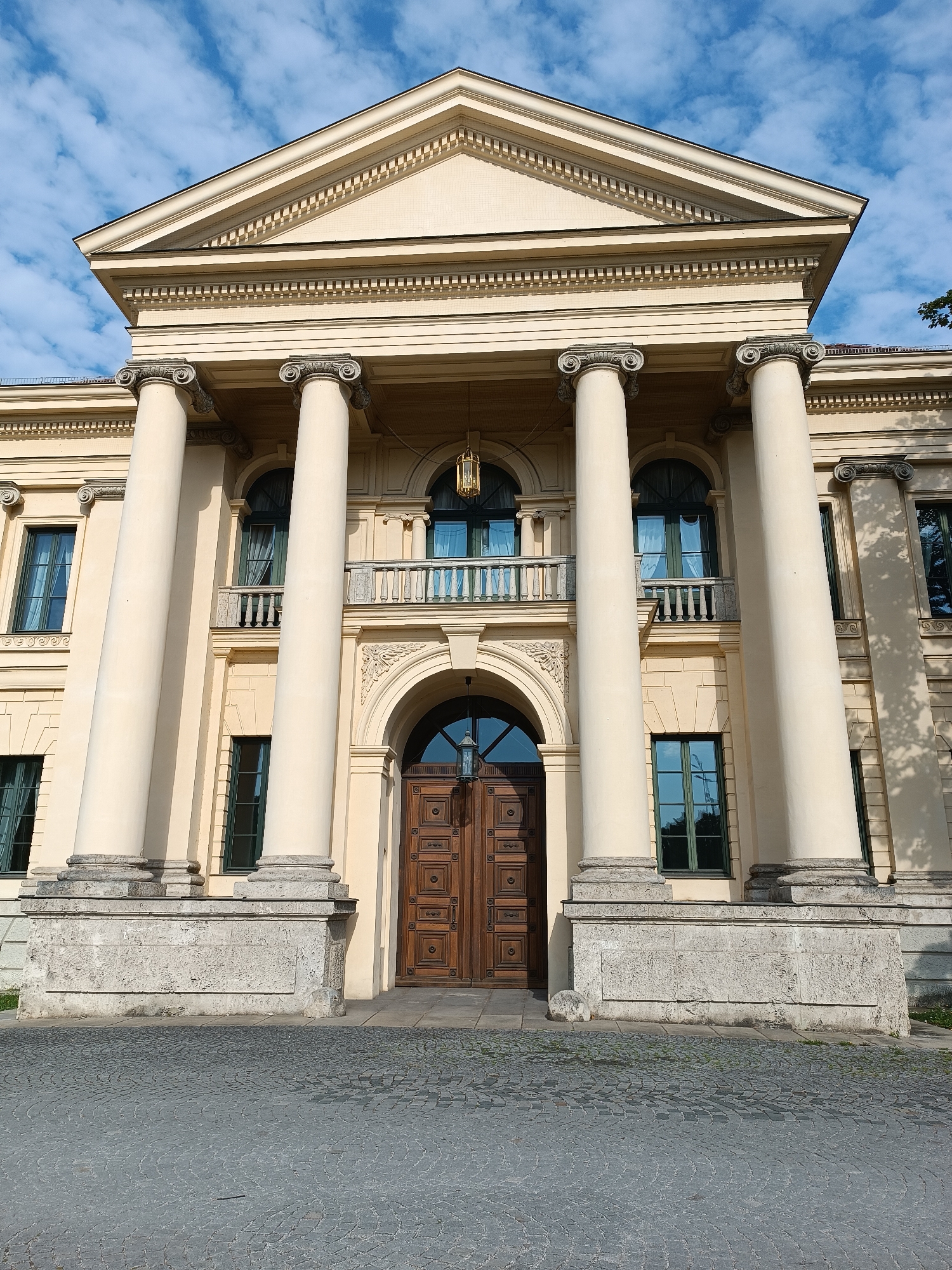 The image shows the Glyptothek museum in Munich, Germany. It is a neoclassical building with a prominent portico supported by Corinthian columns. The facade is made of light yellow stucco and features a triangular pediment. There are several windows on the upper floor, some with balconies. The entrance is a large, ornate wooden door set within an arched entryway. The foreground is paved with cobblestones. The sky is blue with scattered white clouds. There are no people visible in the image.