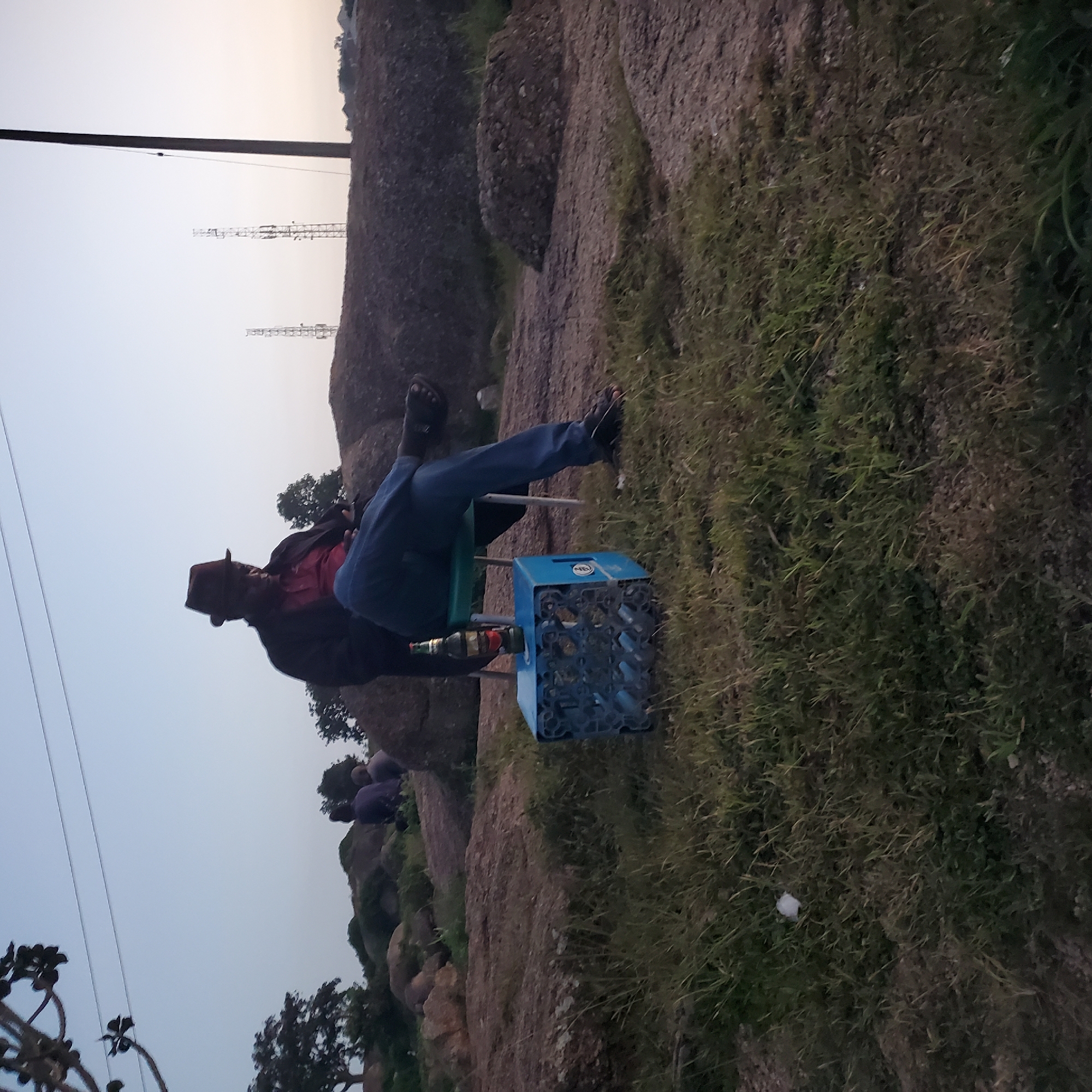 The image depicts an outdoor scene in Jos, Nigeria, at dusk. A man wearing a hat, dark jacket, and blue jeans is seated on a green chair, with his legs crossed. Beside him is a blue crate holding several bottles, and a single bottle of beer rests on top of the crate. The background features rocky terrain, sparse vegetation, and distant structures including utility poles and transmission towers. Two other individuals are partially visible in the distance, seated on a rocky outcrop. The overall environment suggests a somewhat undeveloped or rural area within or on the outskirts of Jos.