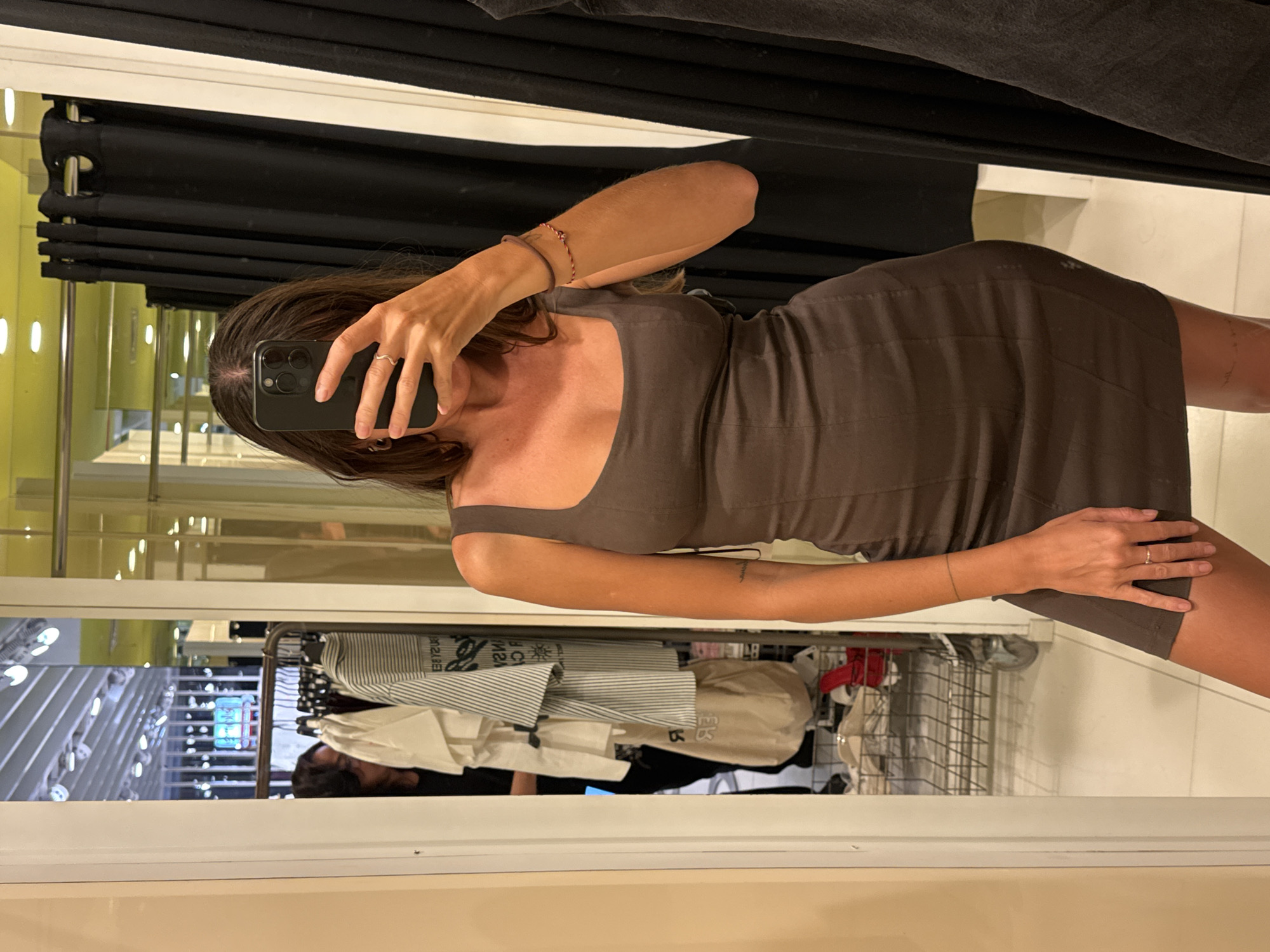 A woman stands in a fitting room with mirrored walls, holding a phone to take a picture of herself. She is wearing a brown dress. The fitting room has a black curtain running along the top. Clothes on hangers are visible through the fitting room's reflection. The scene is likely in a clothing store. There is no information about Kuta, Indonesia in the scene.
