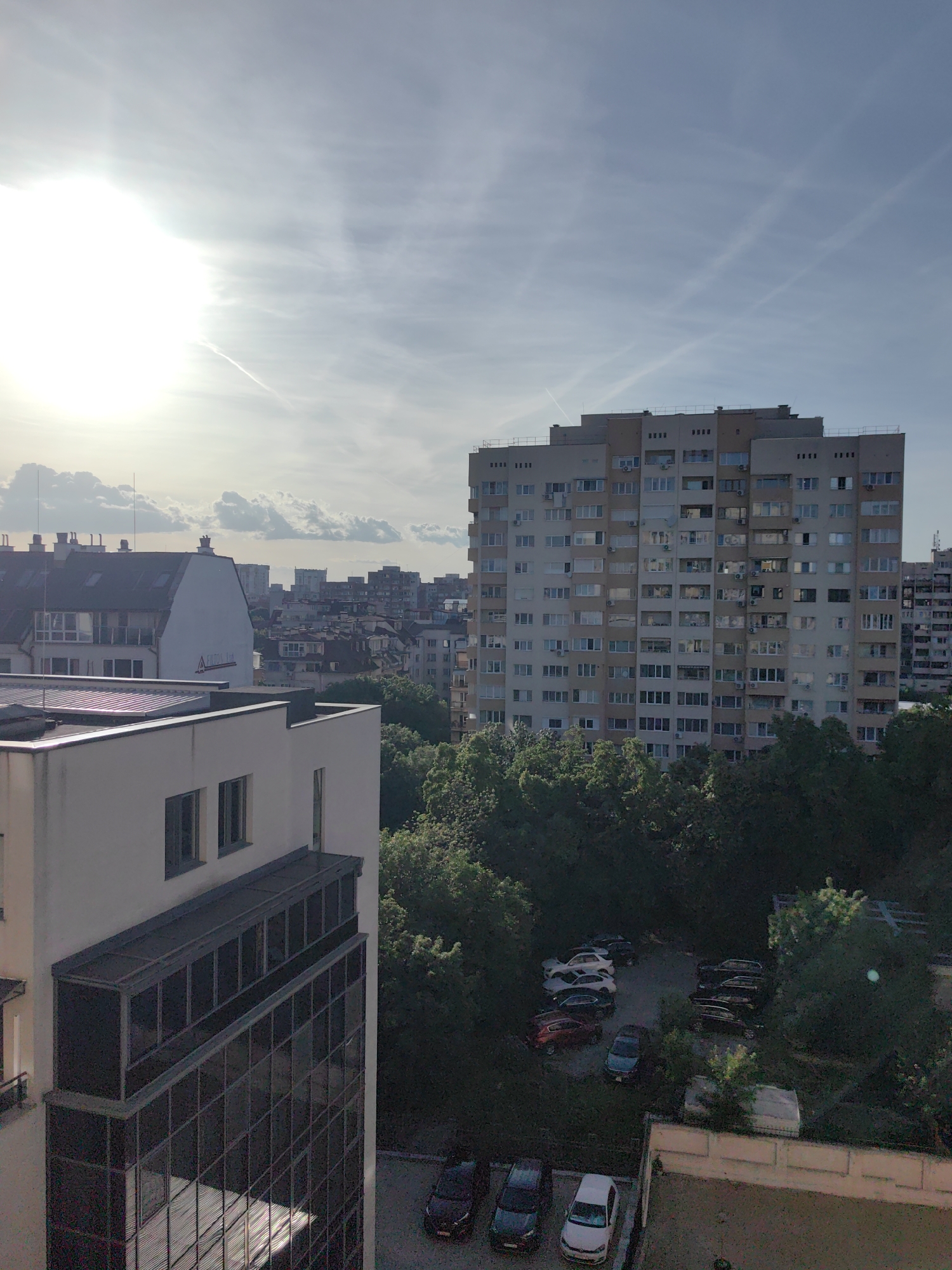 The image shows an urban landscape in Sofia, Bulgaria, featuring a mix of residential and commercial buildings against a partly cloudy sky. The foreground includes parts of a modern, light-colored building with a glass-covered section. Beyond, a high-rise apartment building with light-colored facades dominates the mid-ground, surrounded by lush green trees. A parking area filled with various cars is visible among the trees. In the background, the city's skyline fades into the distance, with multiple buildings of varying sizes and heights. Sunlight is visible on the left, partially obscured by clouds, creating a diffused light across the scene. The sky exhibits contrails, indicating recent aircraft activity.