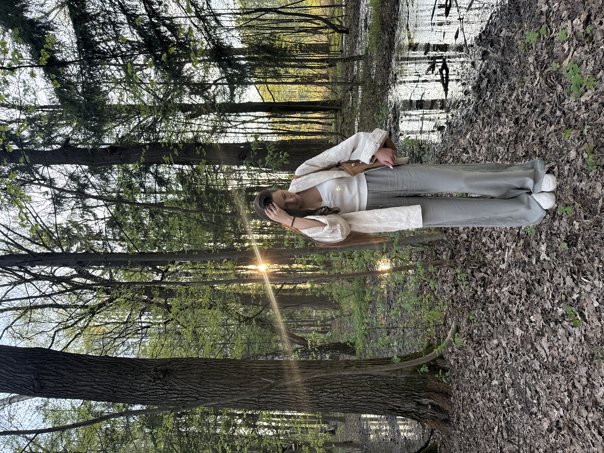 A woman stands in a forest with light filtering through the trees. She wears a white top, a light-colored jacket, and gray pants, with white shoes visible at the bottom of the image. Her right hand is raised toward her hair. The forest floor is covered in leaves, and standing water is visible in the background, reflecting the trees and the sky. The location is in the city of Khimki, Russia.