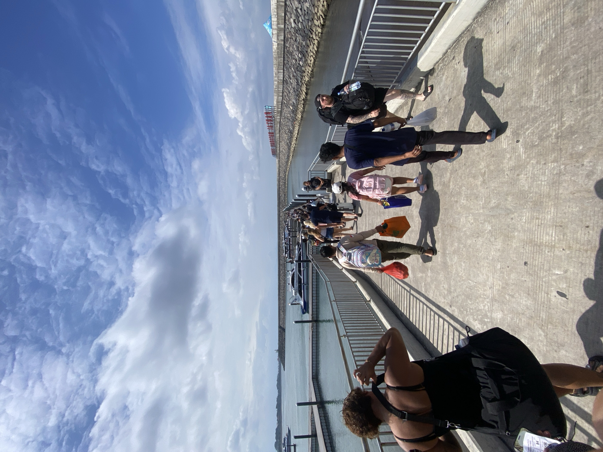 A group of people are walking on a pier in the city of Denpasar, Indonesia. The pier is made of concrete and has a metal railing on the right side. There are boats docked on the left side of the pier, and the water is calm. In the background, there is a long stone wall and a building with red lettering. The sky is blue with some clouds. The people are of various ages and genders, and they are all dressed in casual clothing. Some of them are carrying bags and backpacks. They appear to be heading towards or away from the boats.