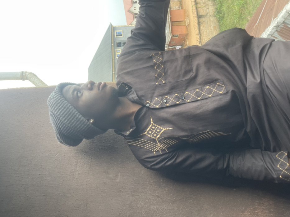 A man wearing a grey beanie and a dark brown long-sleeved shirt with golden embroidery stands against a grey wall. He is looking upwards. Buildings and greenery are visible in the background. The man's shirt features a pocket and decorative stitching around the collar, buttons, and cuffs. The lighting is soft, creating a muted color palette.