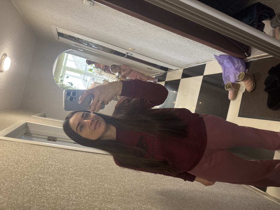 This image depicts an indoor hallway or entryway in the city of Устиново, Russia, captured as a mirror selfie. The primary subject is an adult female with long, dark brown hair, wearing a dark maroon long-sleeved sweatshirt featuring a dark emblem, and light pink trousers. She holds a silver-colored smartphone with a multi-lens camera system in her right hand, with a white nail polish visible. A ring is present on her left ring finger.
Reflected in the mirror, further down the hallway through an arched doorway, a young child with light hair is visible, dressed in a light pink two-piece outfit. The child appears to be looking downwards. Partially visible behind the child is another adult figure, in a reddish garment, facing away from the mirror.
The environment includes walls covered in a textured light beige wallpaper. The floor in the foreground consists of black and white checkered tiles, transitioning to a dark doormat. On the floor near the mirror, a pair of light pink, low-cut, insulated boots and a pair of dark-colored fuzzy slippers are present, alongside a crumpled translucent purple plastic bag. A round, flush-mount light fixture is on the ceiling. Two doors, one white and one dark brown, are visible in the reflection. Beyond the arched doorway, a brightly lit area with what appears to be a window and some green foliage is discernible.