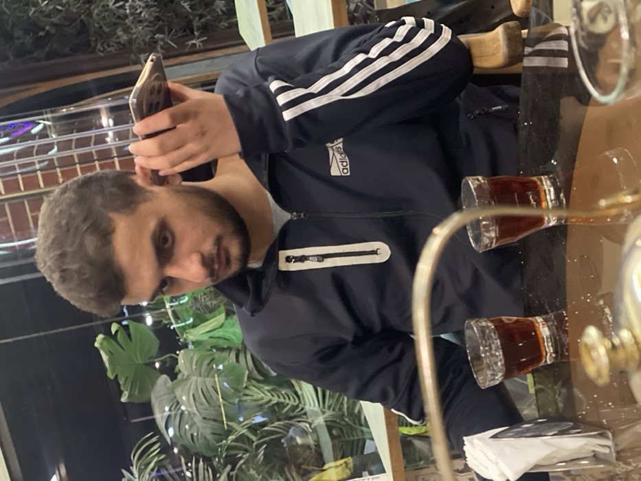 A young adult male, estimated to be in his late teens or early twenties, with short, wavy dark hair and a short beard, is seated at a table. He is wearing a dark blue Adidas track jacket with white stripes on the left sleeve and a white-outlined zippered chest pocket, over a grey t-shirt. He holds a gold-colored smartphone to his left ear with his left hand, appearing to be engaged in a call or listening to audio. His gaze is directed downwards and slightly to his right.
On the dark, reflective table in front of him are two small clear glasses, each containing a dark reddish-brown liquid. A white napkin dispenser is positioned to the left side of the table. A curved metallic object, possibly part of a serving stand, is partially visible in the immediate foreground. The background features dense green foliage, including distinct Monstera leaves, indicating potted plants within an indoor or semi-outdoor setting. Vertical dark elements and subtle reflections of light sources are also present in the background. The lighting appears dim. The specific location within the city of Homs, Syria, cannot be determined from the visual evidence in the image.