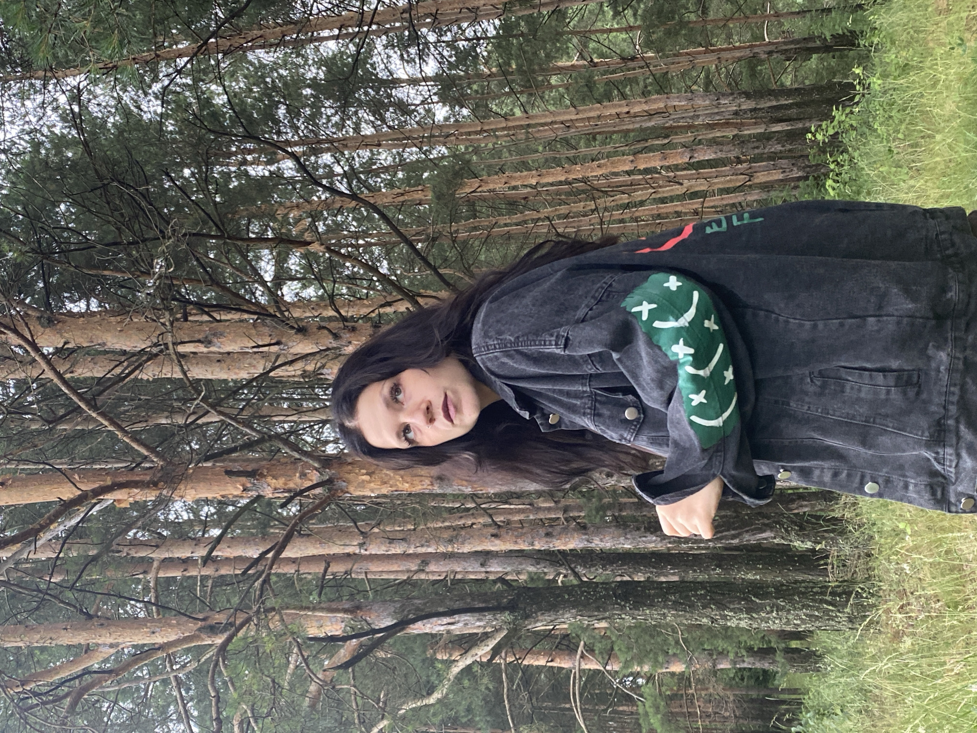 A young woman with long, dark hair stands in a forest of tall, slender pine trees with brown bark and sparse green needles. She is wearing a dark denim jacket with green and red graphic designs on the sleeves. The ground is covered in dry grass and some scattered green foliage. The lighting suggests an overcast day. The woman is looking upwards and to her left, with her hands clasped in front of her. The location is identified as Foki, Russia.