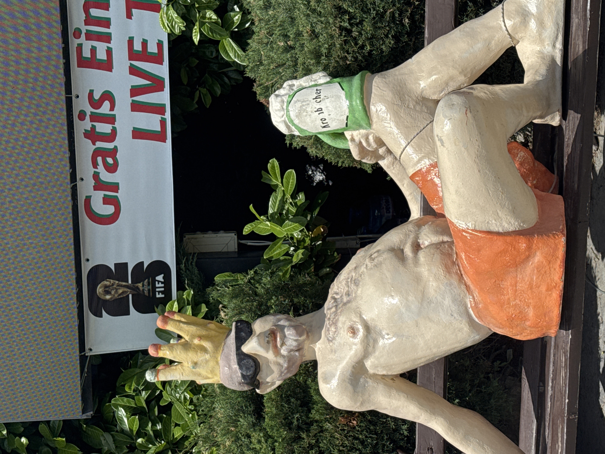 The image shows a statue of a man wearing a crown and sunglasses, sitting on a bench outdoors. He is partially nude and wearing orange shorts. He is holding a green object that resembles a beer mug with white lettering that reads "Kro nb cher." Behind the statue, a banner is visible with text that appears to say "Gratis Ein LIVE T" and a FIFA World Cup trophy logo. The statue and banner are set against a backdrop of lush green bushes and foliage. There is no indication of the city of Varna, Bulgaria in the image itself.