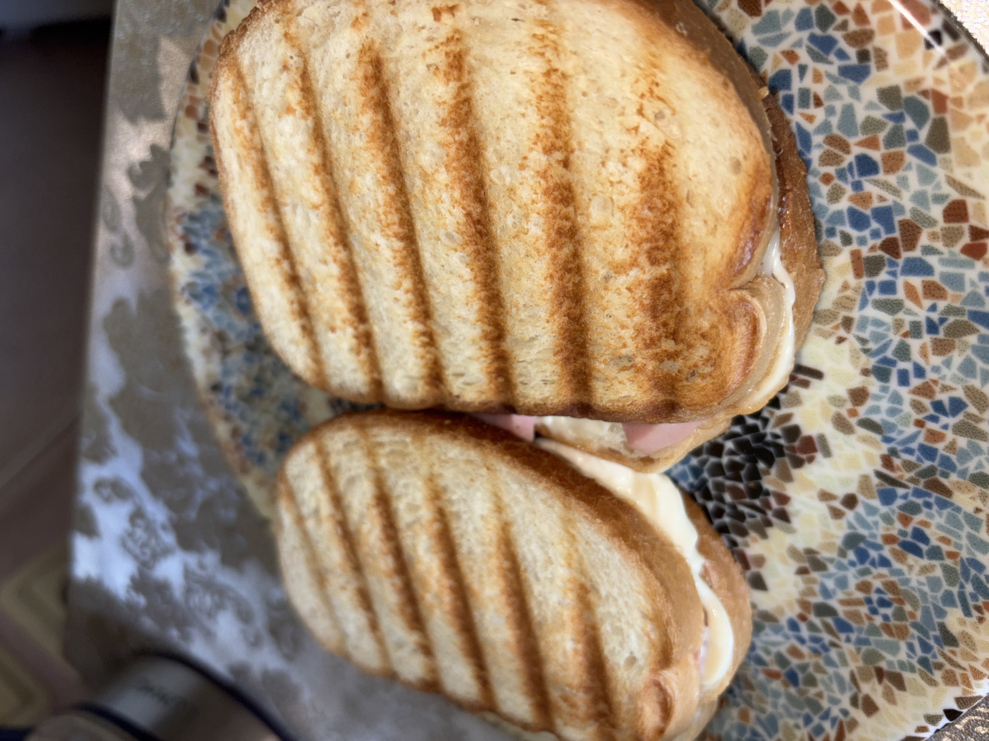 Two toasted sandwiches with grill marks are presented on a colorful mosaic-patterned plate. The sandwiches appear to be filled with melted cheese and ham. The background shows a blurry patterned surface, possibly a tablecloth, and a portion of an appliance on the left. The lighting suggests an indoor setting, likely a kitchen or dining area in a residence within Vsevolozhsk, Russia. There are no people visible in the image.