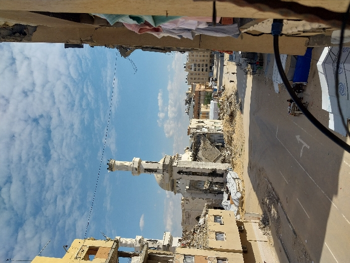An elevated view captures an urban scene of extensive destruction in Khan Yunis, Palestinian Territory, under a blue sky with scattered white clouds. The central focus is a heavily damaged mosque featuring a partially intact white minaret with a circular balcony and dome-like crown, and a partially collapsed main structure with an exposed domed roof. Surrounding the mosque are vast piles of concrete and rebar rubble.
To the immediate left of the mosque, a multi-story building is significantly damaged, with visible structural breaches and debris on its lower levels. A paved road, marked with a white turning arrow, cuts through the lower-center of the frame, separating the immediate foreground from the rubble-strewn areas around the mosque.
In the mid-ground, on the right side of the road, several individuals are visible: a few figures walk in the distance, while closer, a small group congregates around a blue tarp shelter. Another cart, possibly a vendor's, is present near the shelter with a few people around it. In the far distance, other multi-story buildings are visible, some appearing less damaged.
The image is framed from within a damaged structure on the right, where several pieces of laundry (light blue, pink, white) hang from an opening. A dark cable or wire spans across the lower foreground of the image. On the far left, another damaged building partially frames the scene. The overall environment indicates widespread architectural devastation.