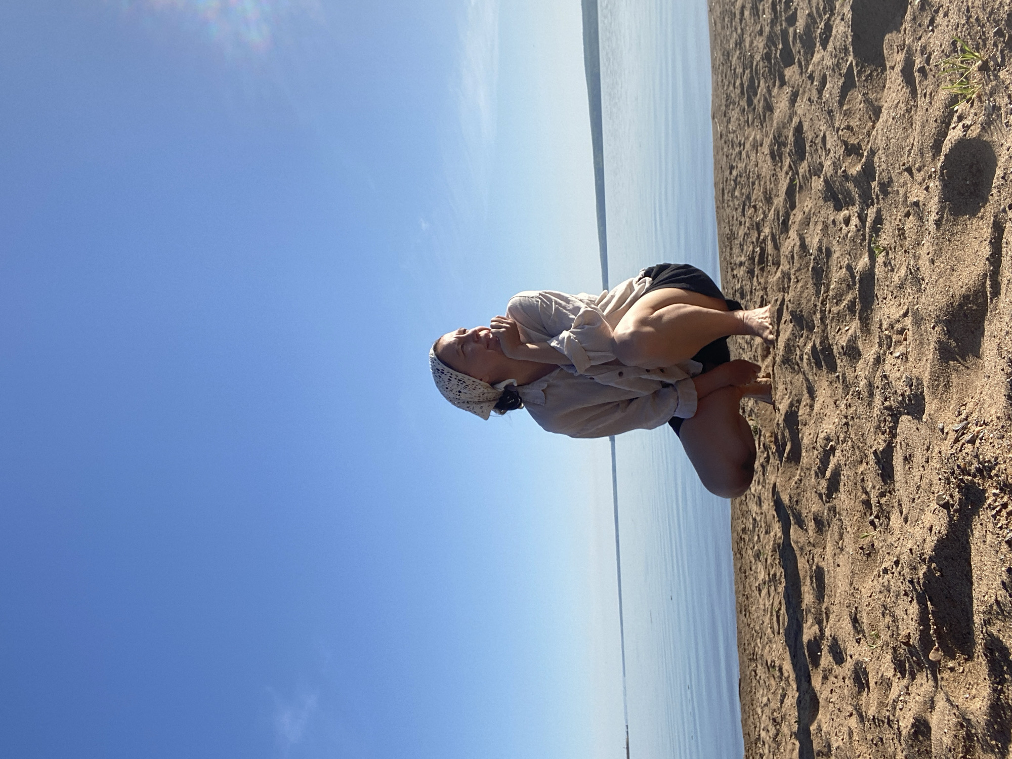 A young woman kneels on a sandy beach alongside a large body of water, likely a lake or reservoir. She is wearing a light-colored shirt, dark shorts, and a patterned headscarf. The woman is positioned between the clear blue sky and the beach, facing towards the water. Her hands are near her face, suggesting she might be laughing or expressing emotion. The scene is bathed in sunlight. The location is the city of Chaikovsky, Russia.