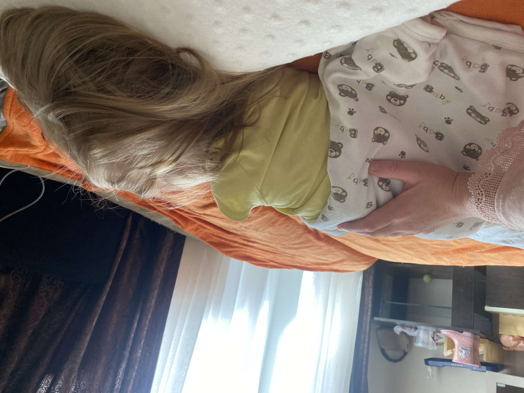 The image depicts an indoor scene within a residential bedroom in Chaikovsky, Russia, during daylight hours. In the foreground, a fair-skinned hand, exhibiting a light-colored garment with a lace cuff at the wrist, rests on a white blanket patterned with brown "Baby" text, bear faces, and paw prints. Beneath this blanket, a person, likely a child, is partially visible, wearing a light yellow top. The child's head, covered with light brown hair, is positioned on a white pillow or cushion. Both are situated on a bed covered with an orange duvet. In the background to the left, a dark-toned bedside table features a pink children's rain boot with a unicorn motif, a clear container, and potentially a small beige bag on its upper surface. A lower shelf of the table holds a small green spherical object. A large window, partially obscured by sheer white and dark patterned brown curtains, provides natural light to the room. A white cable is visible on the orange bedding near the top right.