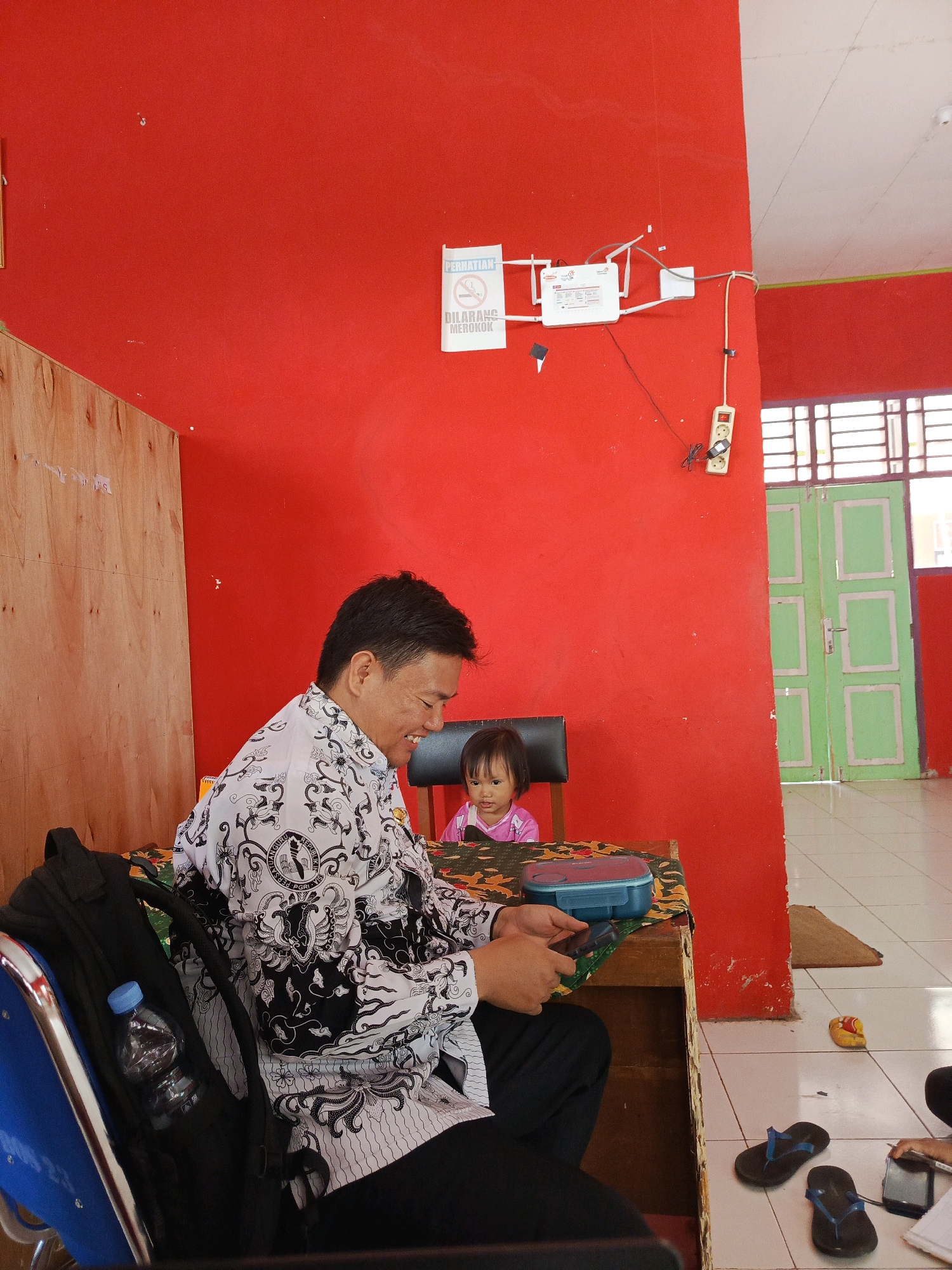 In an indoor setting with vibrant red walls, a man wearing a white shirt with a black and white patterned design is seated at a wooden table, smiling and holding a mobile phone. To his right, a young child in a pink shirt is also seated at the table, looking towards the man. On the table are a blue rectangular container and the man's phone. A black backpack with a water bottle in its side pocket is placed to the left of the man. In the background, a set of light green double doors with glass panes is visible, along with a white ceiling and a white power outlet on the red wall. There are also flip-flops on the tiled floor. The scene suggests a casual or educational environment.