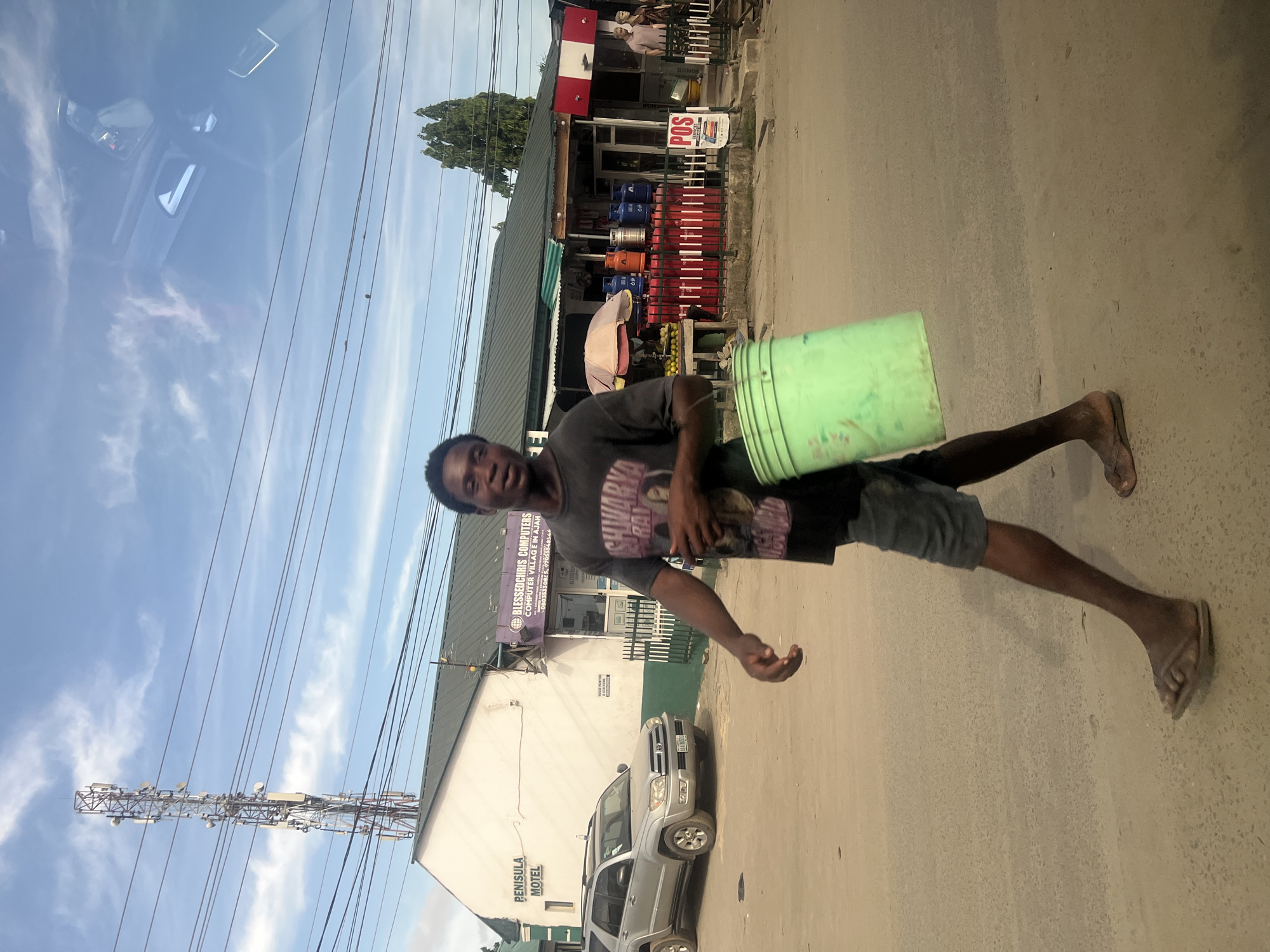 This image, captured from within a vehicle and rotated 90 degrees clockwise from its natural orientation, depicts a bright daylight street scene in Lagos, Nigeria.
In the foreground, a young man, likely of African descent, walks on a paved road, looking towards the camera with a neutral expression. He is wearing a dark grey t-shirt with a partially visible graphic (possibly "MARVEL"), dark shorts, and flip-flops. He carries a light green bucket tucked under his left arm.
The midground reveals a commercial street. A silver Toyota 4Runner SUV is parked on the left, in front of a white building with a green roof. This building features prominent signage, including "PENISULA MOTEL" and "BLESSEDCHRIS COMPUTERS COMPUTER VILLAGE E IN AJAH," along with phone numbers "0803330815" and "07055554544." Further along the street, another establishment displays numerous red, blue, orange, and silver gas cylinders, and a "POS" (Point of Sale) sign. A "BONGOS" sign and market stalls with produce under a beige umbrella are also visible.
The background features a clear blue sky with scattered white clouds. A tall, multi-antenna communication tower stands on the left, with utility lines crisscrossing overhead. Reflections of the vehicle's interior and the sky are visible in the top portion of the frame, indicating the photograph was taken through a car window. The scene portrays a bustling urban environment under clear weather conditions.