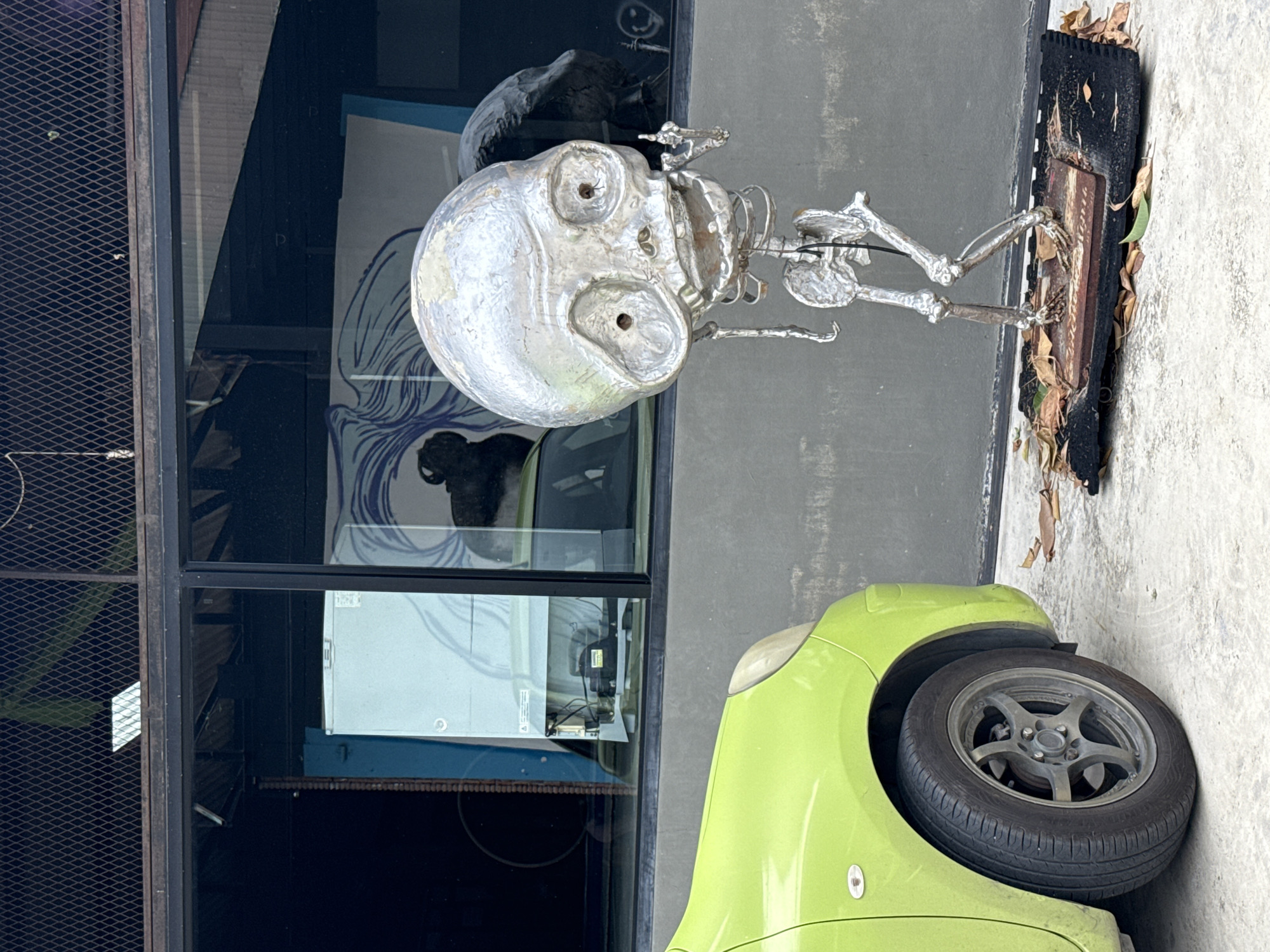A metallic sculpture resembling a skeleton with a large skull is positioned on a black rectangular base adorned with dried leaves on a concrete surface. The sculpture is in the foreground to the right. To the left, the front portion of a bright green car is visible, showing its front tire and fender. Behind the sculpture and the car is a building with a gray concrete wall and large windows. Reflections of objects, including a white appliance and a mural with blue wavy lines, are visible in the windows. A dark metal grate is present above the windows. The scene appears to be outdoors, possibly in an urban or commercial setting in the Ko Samui District, Thailand.
