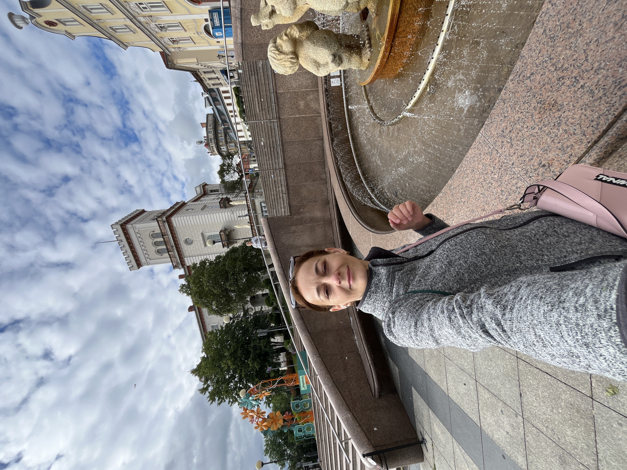 In this wide-angle, eye-level selfie, a woman with brown hair pulled back and wearing sunglasses on her head and a grey zip-up jacket stands in a paved outdoor area. She has a pink purse slung over her shoulder. To her left, a large, multi-tiered staircase leads up to buildings, with decorative floral structures and large letters spelling "BBB" visible at the base of the stairs. To her right, a circular fountain with water jets and small statue figures is visible. In the background, various buildings with architectural details and a cloudy sky with scattered blue patches are present. The scene appears to be a public plaza or park area within a city.