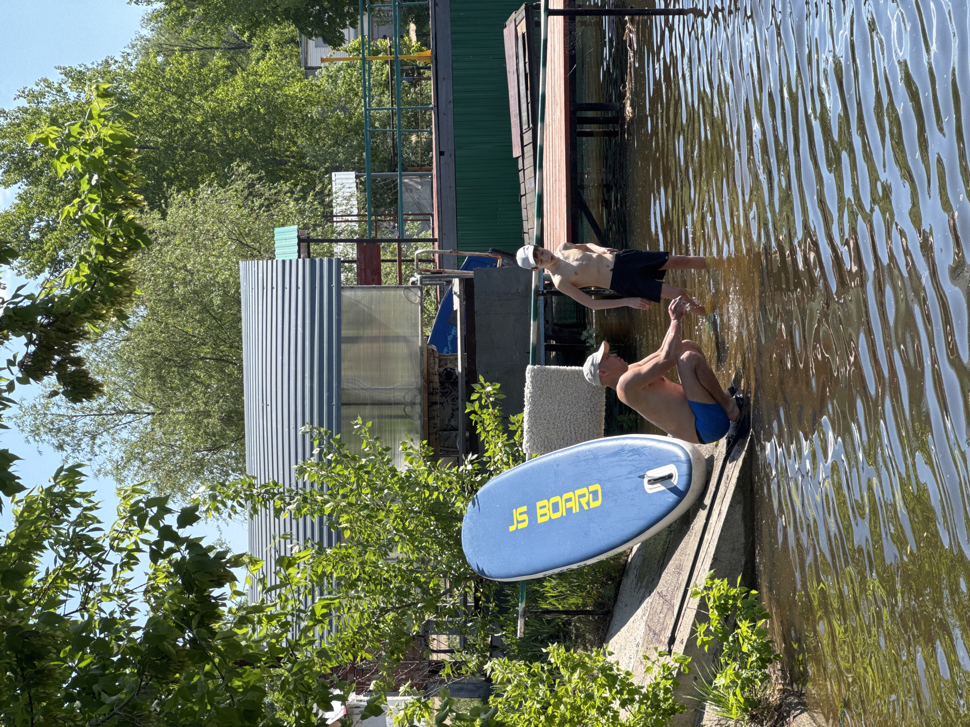 Two shirtless males are near the water’s edge, presumably in Chaikovsky, Russia. One male wears a white cap, black shorts, and stands in the water. The other male wears a white cap and blue swimming trunks and is crouching by the edge. A blue paddleboard labeled "JS BOARD" is adjacent to him. The scene features a mix of natural elements (water, trees) and man-made structures, including a corrugated metal building and a wooden dock.