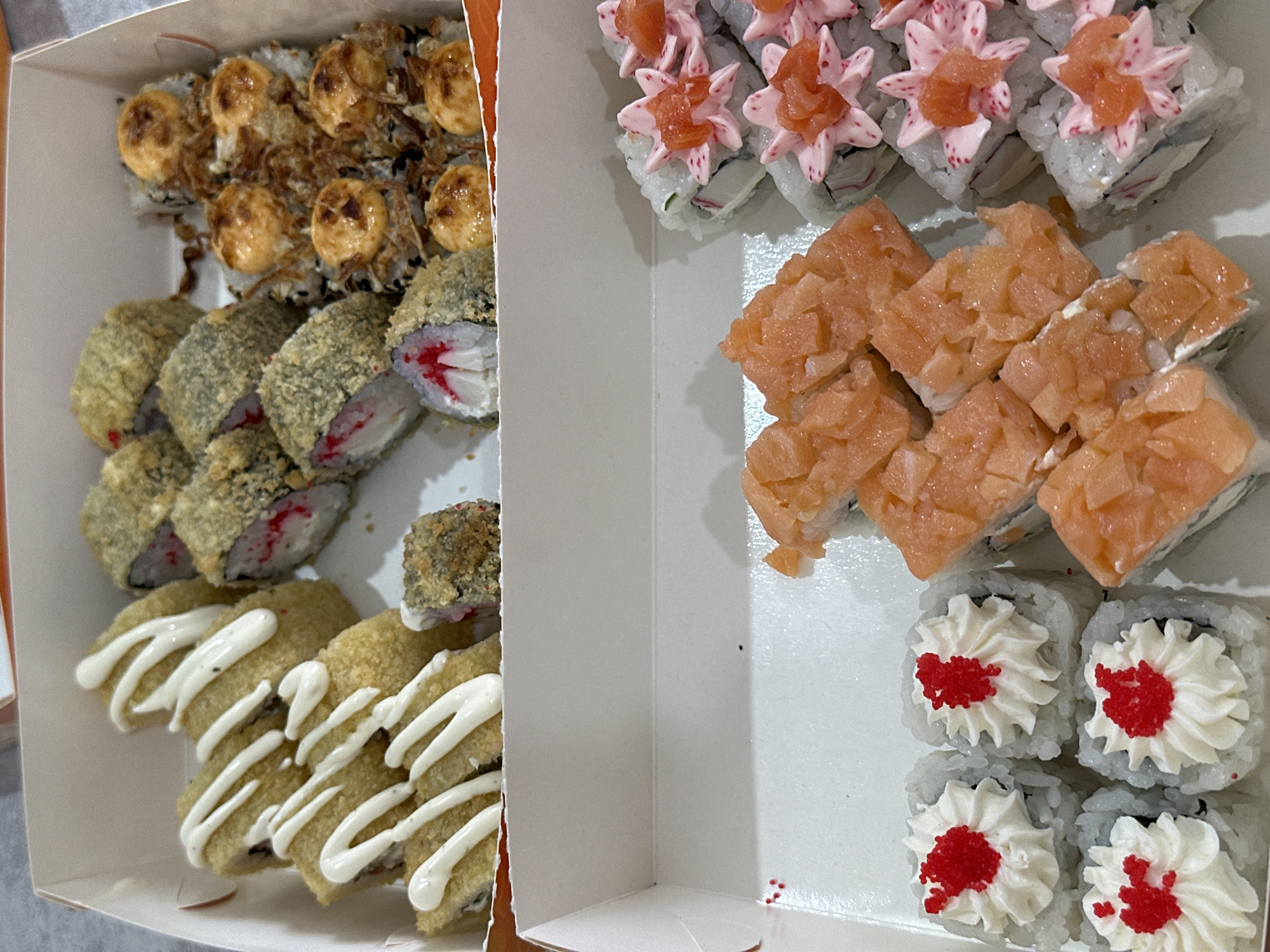 This media file is a top-down shot of an open white cardboard container filled with various types of sushi. The container is divided into two sections.
The left section contains several types of fried sushi rolls. Some are topped with a golden-brown, creamy sauce and crispy fried onions. Others are coated in a golden-brown batter and appear to have a filling with red elements, possibly imitation crab. The bottom of this section features several rolls drizzled with white sauce, likely mayonnaise.
The right section of the container holds different varieties of sushi. The top row shows rolls with a light pink creamy topping and small pieces of salmon, arranged to resemble flower petals. Below these are several rolls topped with diced raw salmon. The bottom row contains four sushi rolls topped with white cream cheese, arranged in a swirl pattern and decorated with red tobiko (flying fish roe). A few loose red roe beads are visible in the bottom right corner of the container.
The lighting suggests an indoor setting, likely a home or a restaurant, with no specific time of day or weather cues. The focus is entirely on the food, showcasing a diverse selection of sushi presented in a visually appealing manner. There is no visible text in the image. The overall impression is one of a ready-to-eat meal or takeout order.