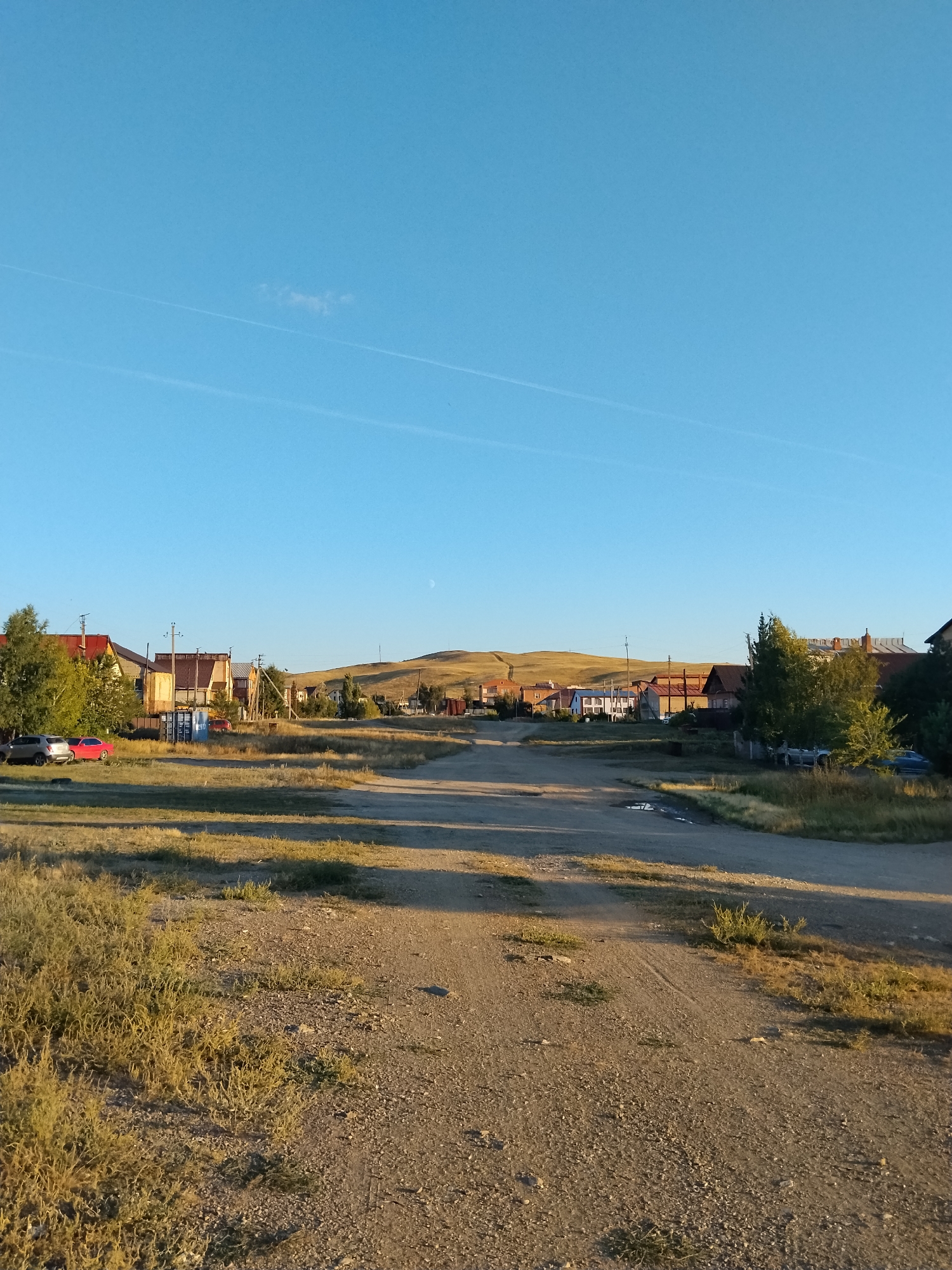 The image depicts a rural village scene under a clear blue sky with thin contrails. The foreground is a dirt road with sparse dry grass and gravel. In the middle ground, a row of houses with pitched roofs and varying architectural styles is visible, some with red roofing. A few trees, including one with green foliage, are scattered among the houses. Several cars, including a silver SUV and a red sedan, are parked on the left side of the road. In the background, rolling hills are covered in dry grass, and a faint crescent moon is visible in the sky. The overall impression is one of a quiet, rural settlement bathed in the warm light of what appears to be late afternoon or early morning. The specific location within Kokshetau, Kazakhstan cannot be determined from the image alone.