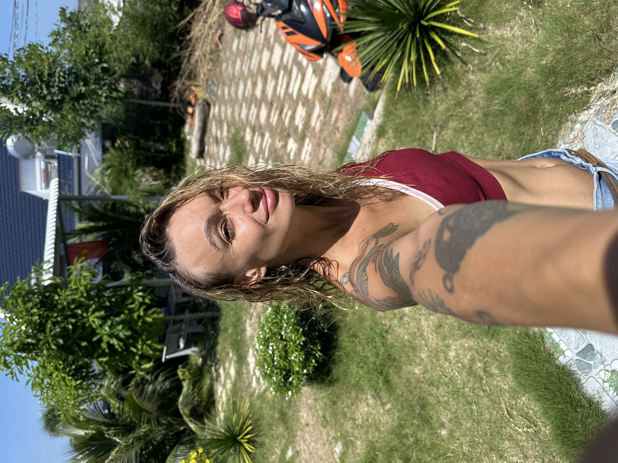 The image depicts a woman in Mũi Né, Vietnam, taking a selfie outdoors. She is positioned in the foreground, with her arm extended towards the camera. The woman has blonde, wet hair and is wearing a maroon sports bra and denim shorts. Tattoos are visible on her left arm and shoulder. Behind her, a paved pathway leads to a residential building with a dark tiled roof and a flag hanging from an overhang. The environment is characterized by lush green grass, palm trees, and other tropical vegetation. In the background, an orange and black motorcycle with a red helmet on its seat is parked. The scene suggests a sunny day in a tropical or subtropical climate.