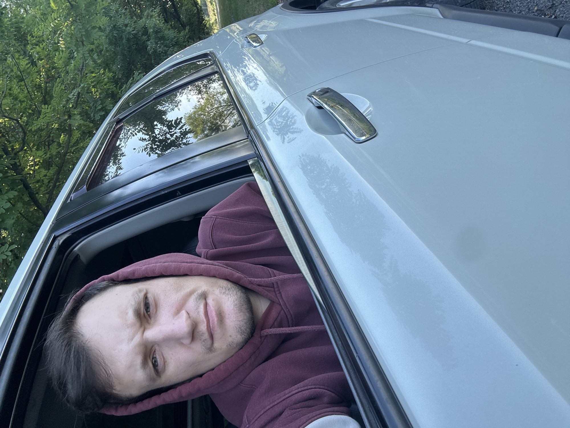 A medium shot captures a man wearing a maroon hoodie, his face partially obscured by the hood, looking directly at the camera from inside a car. He is seated in the driver's seat, with the car door open and the window rolled down. The man has short, dark hair, a light complexion, and a faint stubble on his face. His expression is neutral. The car's interior is visible in the background, with the black dashboard and the car's light-colored exterior dominating the foreground. The car is parked outdoors, with lush green trees and foliage visible through the open window and in the reflection on the car's surface. Sunlight filters through the leaves, casting dappled shadows. The image appears to have been taken during daylight hours. There is no visible indication of the specific location in the city of Makiv, Ukraine.