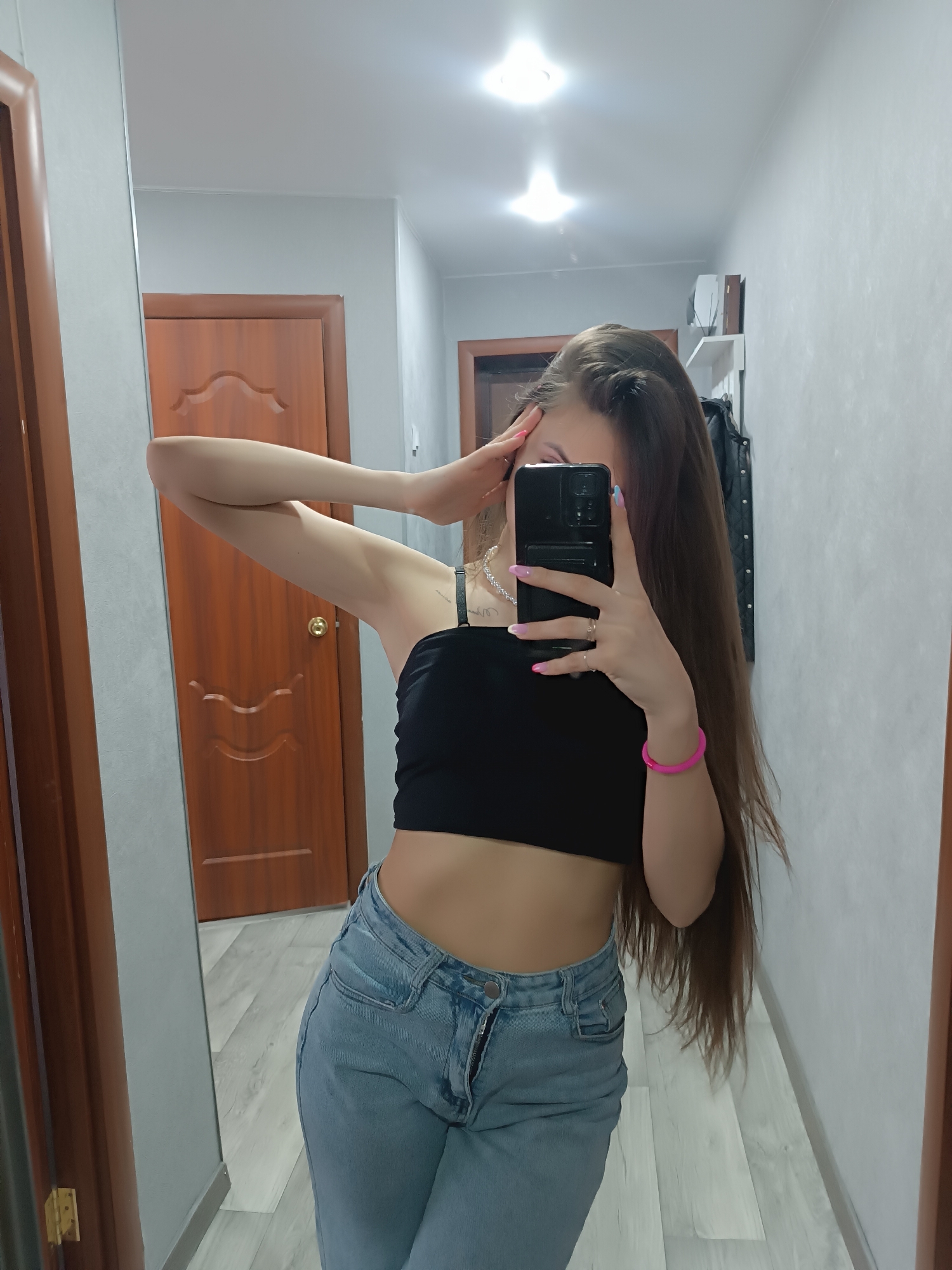 A woman is seen taking a selfie in a mirror. She is wearing a black crop top and light blue jeans. Her long brown hair is partially covering her face. She has a pink bracelet on her right wrist and a necklace around her neck. She is holding a black smartphone with pink painted nails. The location appears to be a hallway in a house or apartment, with wooden doors and light gray walls. There is a shelf with items on the wall in the background.