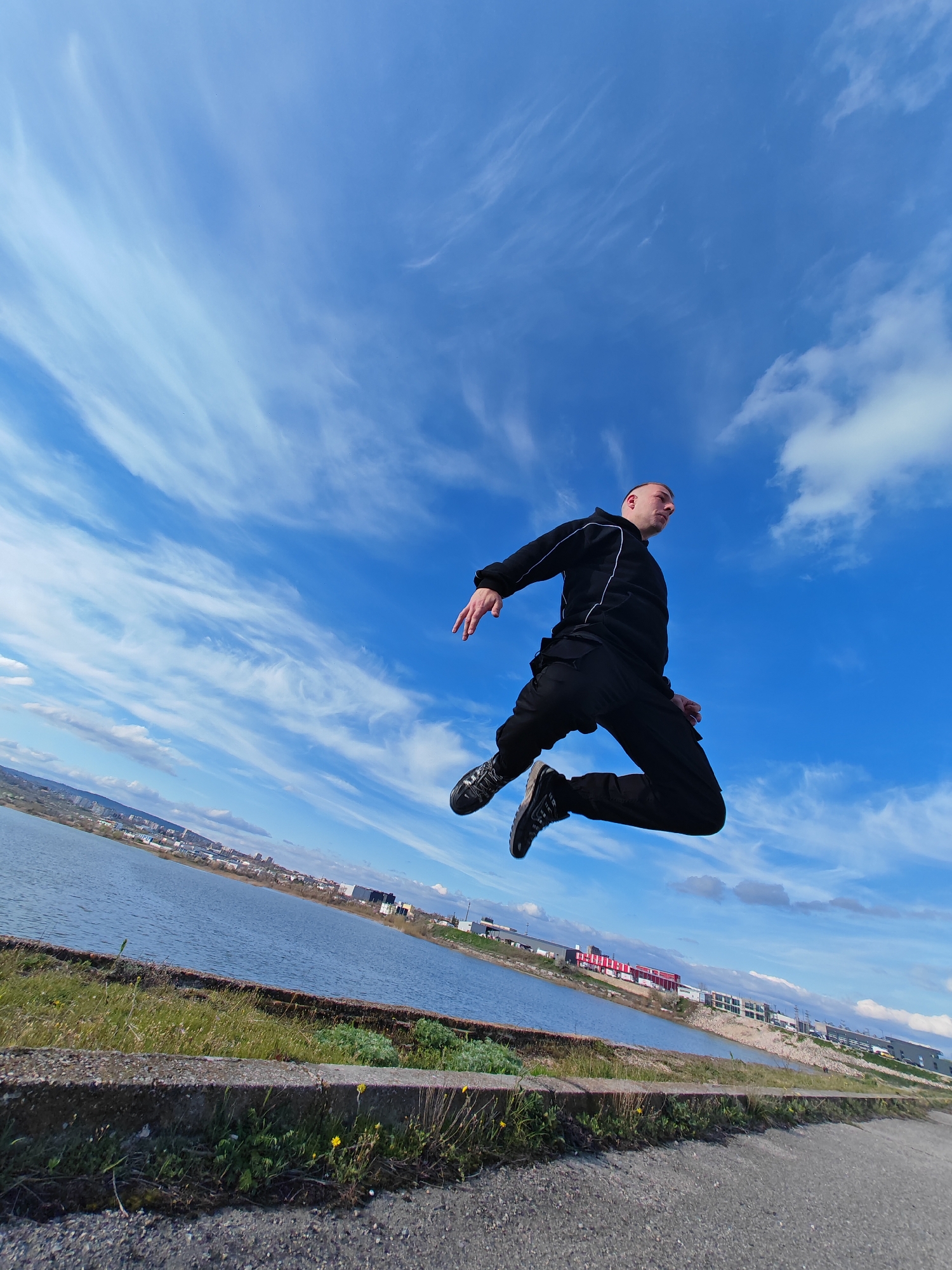 A man in a black tracksuit is captured mid-air in a dynamic jump against a backdrop of a bright blue sky with wispy white clouds. He is positioned to the right of the frame, with his body angled slightly upwards. The land below consists of a concrete walkway, a grassy verge, and then a body of water. Beyond the water, there is an urban landscape with buildings and distant hills. Based on the distinctive industrial architecture, cityscape layout, and body of water to the left, this location is near the industrial area on the outskirts of Shumen, Bulgaria. The man's action suggests athleticism or recreation, contrasted by the utilitarian environment.