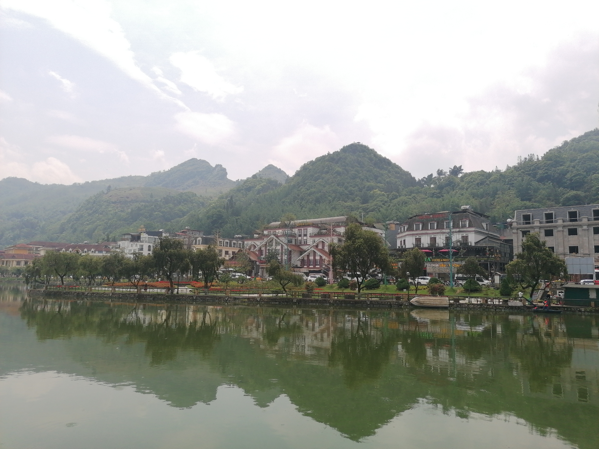 The photograph captures a panoramic view of Sa Pa Ward, Vietnam, showcasing a tranquil body of water in the foreground reflecting the cityscape and the mountainous backdrop. The water surface is calm, displaying a green hue. A paved walkway lined with trees and a low barrier runs along the waterfront.
Behind the walkway, a row of buildings varying in style and height are visible. Some buildings have distinct architectural features such as steeply pitched roofs and dormer windows. Lush green hills and mountains form the background, their peaks partially obscured by cloud cover. The sky is overcast, contributing to a soft, diffused lighting across the scene.
There is a mix of trees, buildings, and natural elements. There is no significant human activity visible in the scene.