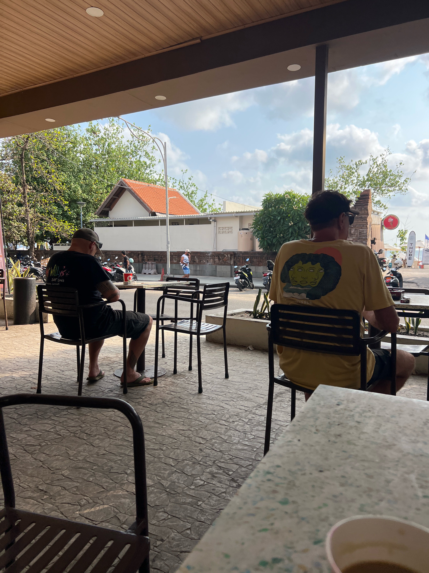In Kuta, Indonesia, two men are seated outdoors at tables on a paved patio beneath a covered overhang. The man on the left, wearing a black t-shirt and a baseball cap, is facing away from the camera, looking at a table. He is wearing shorts and flip-flops, with tattoos visible on his legs. The man on the right, also facing away, is wearing a yellow t-shirt with a graphic design of faces and a sun. He is wearing sunglasses and shorts. In the background, several motorcycles are parked, and people are walking along a street. Buildings with red tile roofs and white walls are visible, along with trees and a bright sky with clouds. The scene appears to be a casual outdoor dining or resting area.
