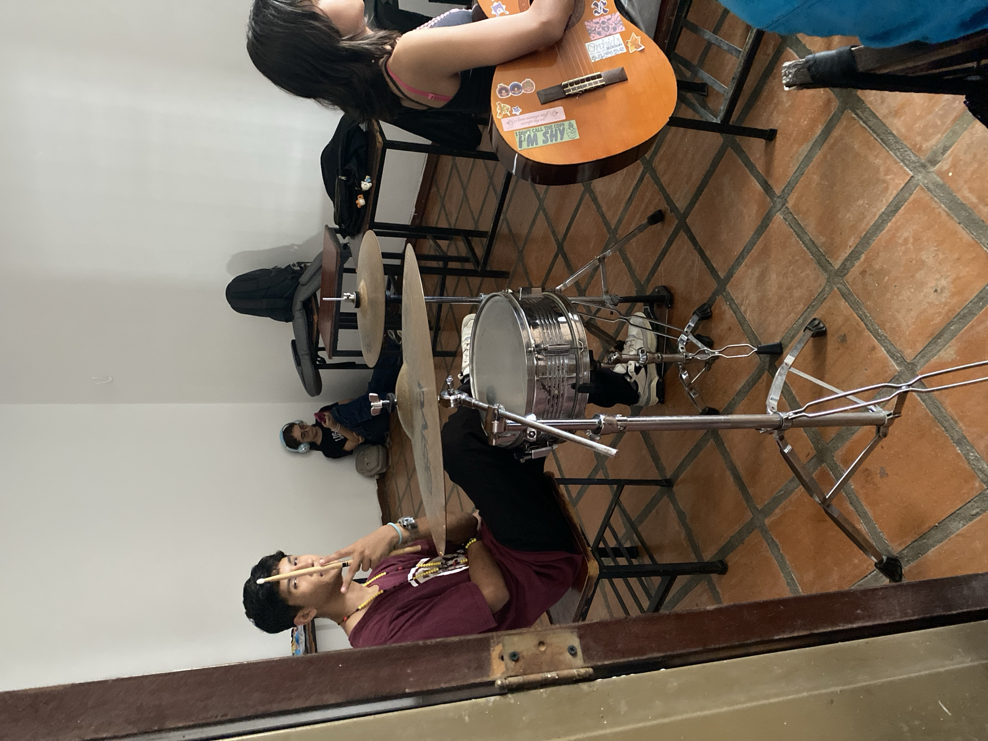 The scene appears to be indoors, likely in a music room or practice space, potentially within a residence in Tinaquillo, Venezuela. The floor is tiled with red-brown, square tiles arranged in a diamond pattern.
A young man is seated at a drum set, holding drumsticks and preparing to play. He is wearing a maroon t-shirt, dark pants, and a beaded necklace. The drum set includes a snare drum, a cymbal, and associated stands. The snare drum is silver, and the stands are made of chrome.
Behind the drums, a young woman is seated with a guitar. The guitar is light brown with several stickers on it, including one that reads "I DON'T CALL THE COPS". She has long, dark hair and is wearing a black tank top.
In the background, another person is lying down, wearing a black t-shirt, jeans, and headphones. There is a black backpack on a chair near them.
The room is otherwise sparsely furnished. The scene is captured from a slightly elevated perspective, possibly through a doorway or from a higher viewpoint.