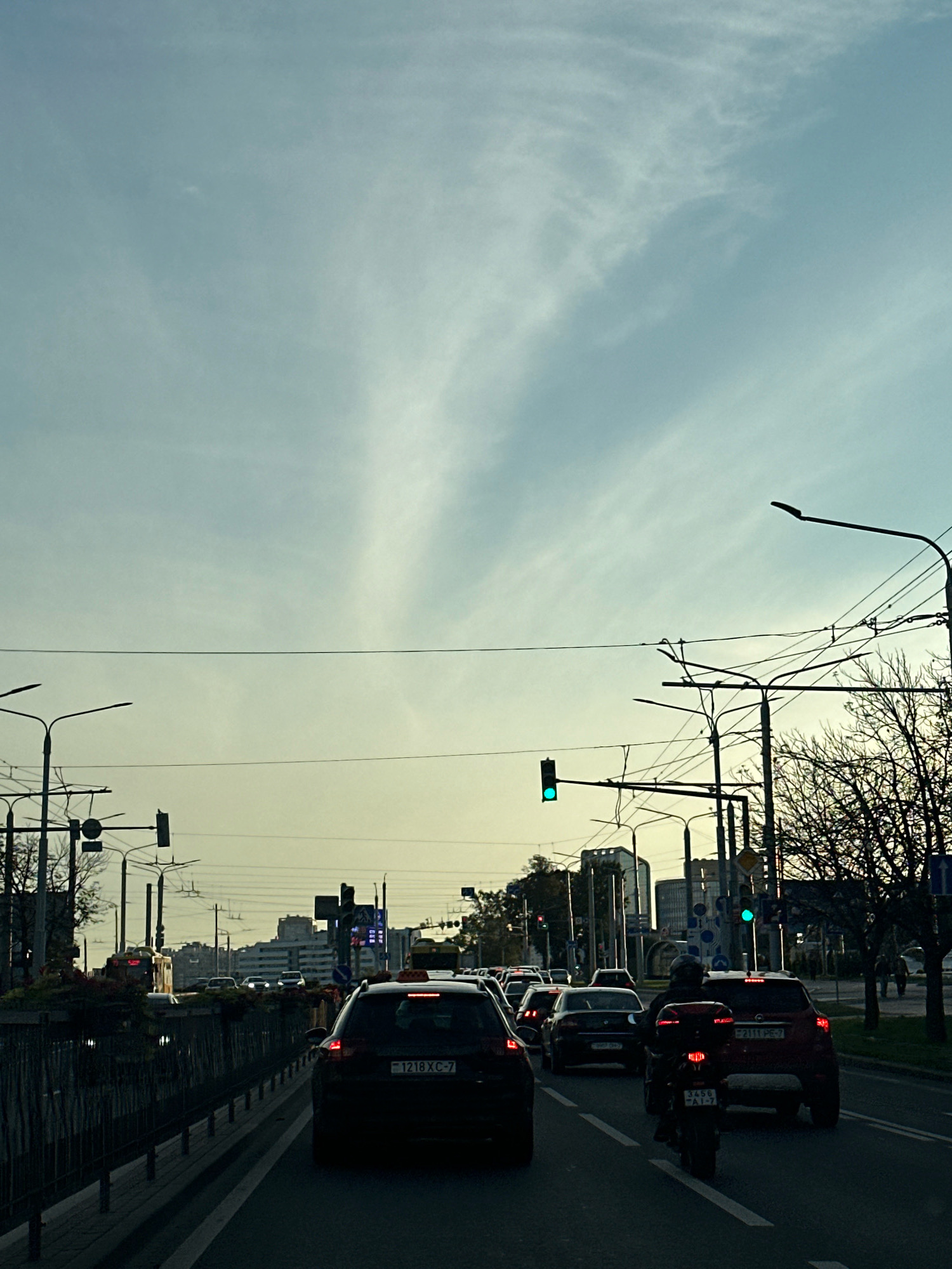 The image depicts a traffic scene in Minsk, Belarus, likely on a city street during daylight. The sky is a pale blue with wispy clouds, transitioning to a yellowish hue near the horizon, suggesting either early morning or late afternoon. Several cars and a motorcycle are visible on a multi-lane road, indicating moderate traffic congestion. Traffic lights are present, with at least one showing a green signal. Lampposts and overhead wires are prominent features of the urban infrastructure. In the background, buildings of varying heights, including what appear to be apartment blocks and commercial structures, form the cityscape. Trees with sparse foliage are also scattered throughout the scene, some with bare branches. A dark metal fence runs along the left side of the road, bordering a sidewalk or pedestrian area. The overall atmosphere is that of a typical urban commute.