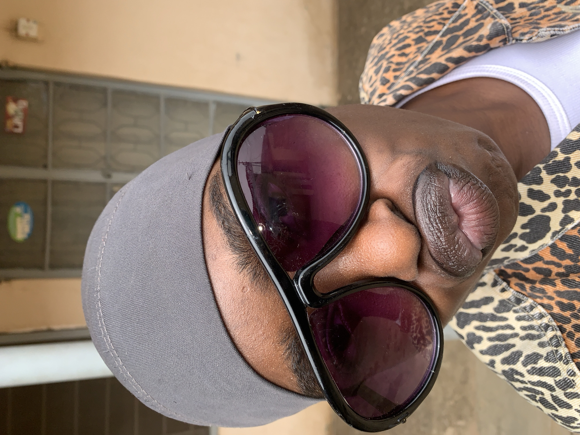 Here is a description of the image:
An eye-level close-up portrait shows a person wearing a grey cap, large black-rimmed sunglasses with purple lenses, a white undershirt, and a leopard-print jacket. The background consists of a light-colored wall with a door and window. The location is identified as Rafin Par, Nigeria.