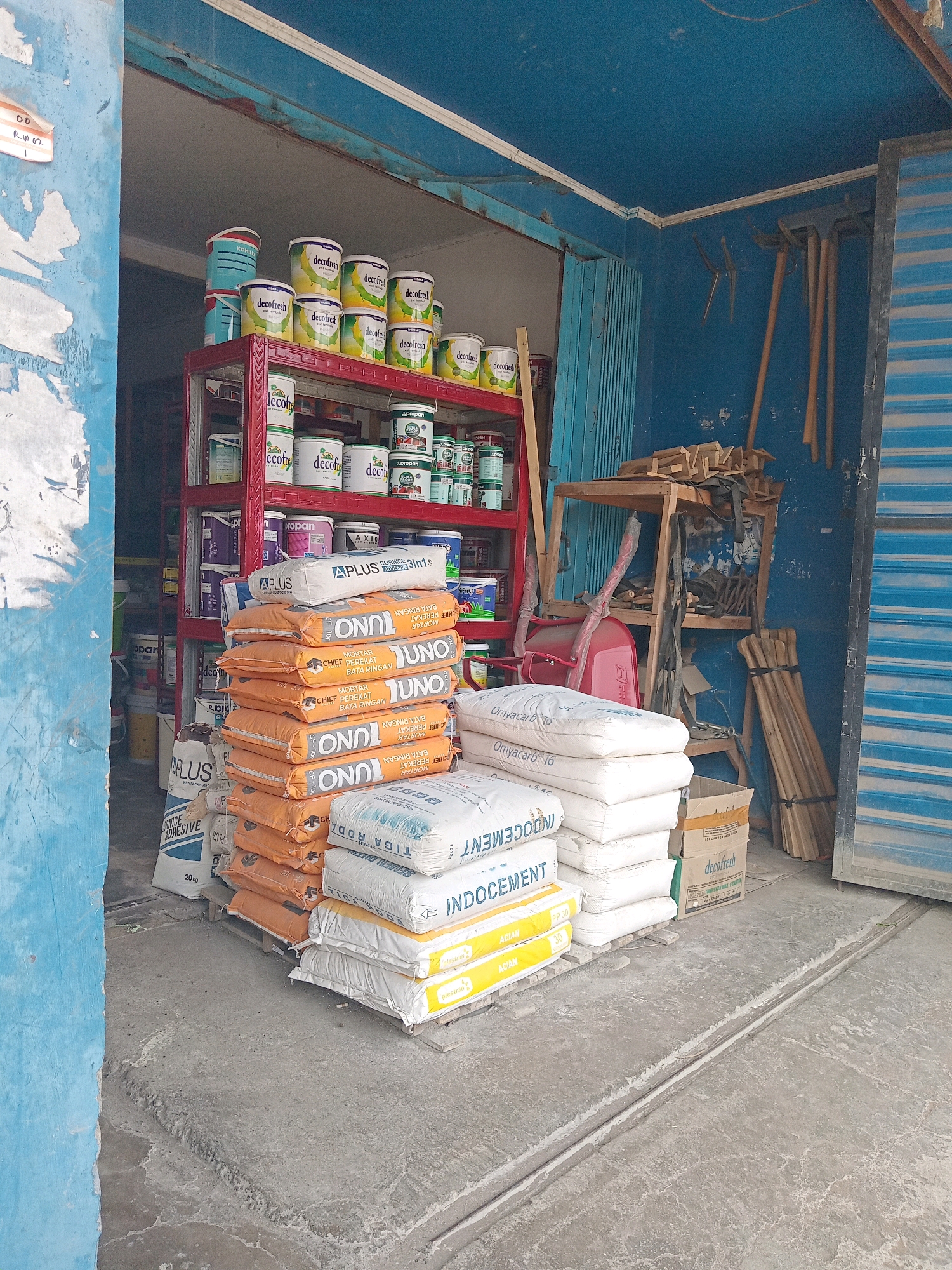 In Sukorejo Kulon, Indonesia, this image captures a storefront with an open facade displaying various construction and home improvement supplies. The interior is painted a vibrant blue, with some signs of wear and tear on the walls. A red metal shelving unit is filled with cans of paint, predominantly labeled "decofresh" in yellow and green, and other unlabeled cans in shades of green and purple. On the floor, a stack of orange bags, each marked with "JUNO MORTAR" and "CHEF PERAKAT BATA RINGAN," is prominently displayed. To the right of these are white bags of what appears to be cement or adhesive, with labels like "A PLUS," "INDO CEMENT," and "OMYACARB." Further to the right, leaning against the blue wall, are several gardening tools, including rakes and what look like hoes, along with a red wheelbarrow and a wooden stand holding more tools and supplies. A blue and white striped metal shutter partially closes off the right side of the storefront. The ground is a concrete floor, showing some rough patches and a visible drainage channel.