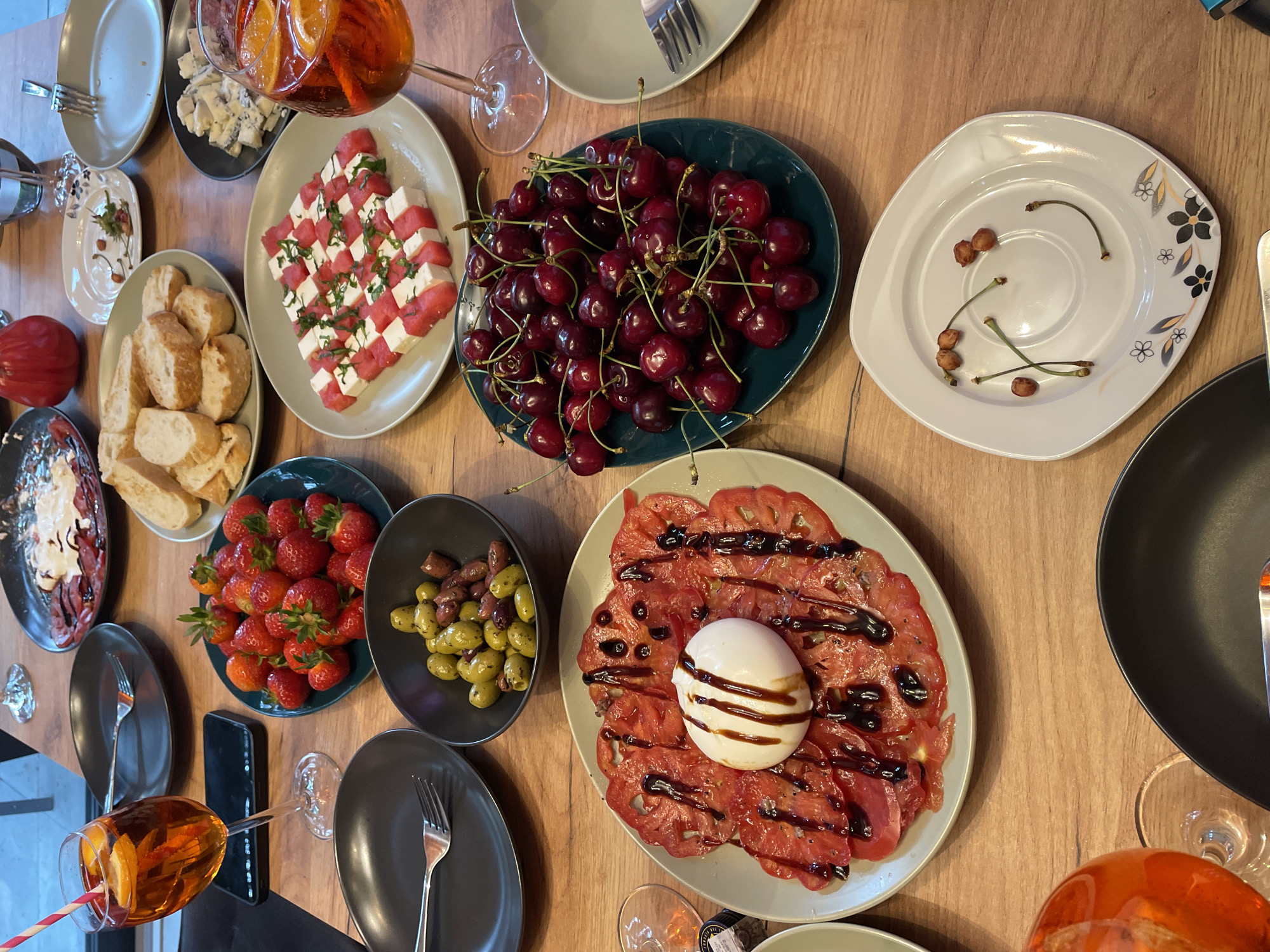 A high-angle, close-up shot captures a wooden table laden with various dishes and drinks, suggesting a meal or gathering. In the foreground, a black plate with a fork sits to the left, and a black bowl holding olives and what appears to be some chopped prosciutto to the right. Further back, a large platter holds thinly sliced tomatoes topped with a ball of burrata cheese and drizzled with balsamic glaze. To the right of the burrata dish, a dark green plate is piled high with fresh cherries.
In the upper left quadrant, a stemmed glass filled with an orange aperitif cocktail, garnished with an orange slice and a straw, is prominent. Next to it, a black smartphone rests on the table. A plate with sliced bread and a red object, possibly a tomato or pepper, sits above the bread. Further right, a plate displays cubes of watermelon and feta cheese, sprinkled with fresh herbs. Another stemmed glass with a similar orange cocktail is visible near the watermelon and feta dish.
A plate of strawberries is positioned between the aperitif cocktail and the watermelon and feta. In the upper portion of the image, a black plate contains what looks like carpaccio with a creamy white sauce. There are several other plates and glasses scattered across the table, contributing to a sense of abundance and a shared meal. The overall setting appears to be indoors, with diffused lighting. The scene is static, with no visible people or explicit actions taking place.