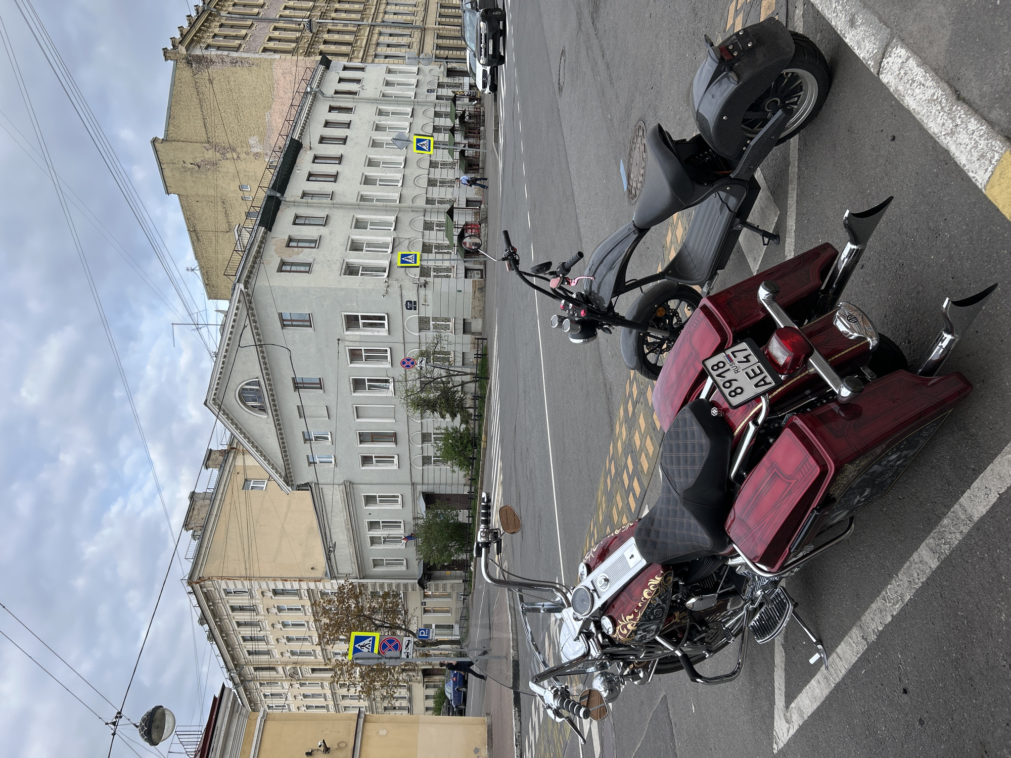The image shows two motorcycles parked in a city street. A burgundy and chrome motorcycle is positioned to the lower right. A black scooter is parked behind it. The street has white lane markings and yellow hatched markings indicating a no-parking zone. Buildings line the street, appearing to be residential and commercial structures, varying in height and color. There are pedestrian crossing signs visible. The sky is overcast. The motorcycle license plate indicates it is in Saint Petersburg, Russia. A few pedestrians and cars are visible in the background.