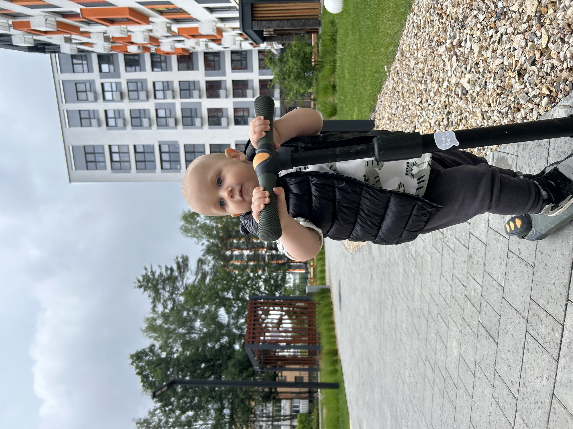 An outdoor medium shot captures a young toddler boy standing on a scooter on a paved walkway. The boy is wearing a black puffer vest over a white t-shirt with a grey and black animal print, and grey sweatpants. He has light skin and blonde hair, with blue eyes looking to the left of the frame. The scooter's handlebars are green with an orange accent, and a sticker with Russian text is visible on the scooter's pole. The background features a modern apartment complex with white and orange facades, green grass, trees, and a cloudy sky. The scene is set in a developed urban environment.