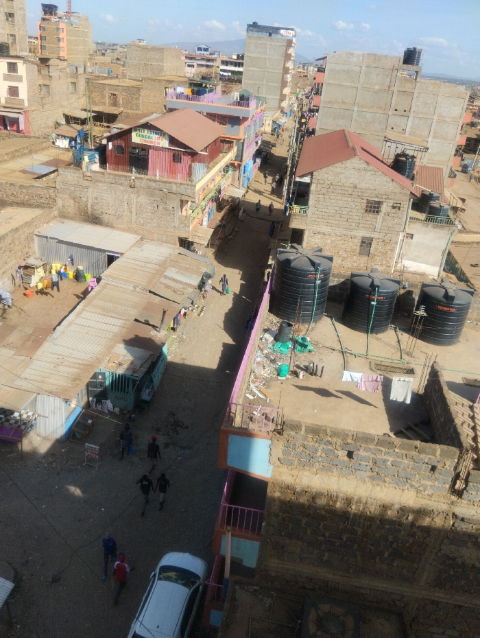 This aerial image depicts a densely populated urban environment within Witeithie ward, Kenya. The scene is an overhead view of a narrow, unpaved dirt street flanked by numerous multi-story buildings and single-story structures.
**Buildings and Structures:** Most buildings are constructed from concrete blocks, exhibiting varying states of finish from exposed stone to painted facades. Many feature corrugated iron roofs, some reddish-brown, others silvery-grey. Visible businesses include "HOLY TRINITY GLOBAL CHURCH" with a brown roof and a structure labeled "GANGAS." Several black plastic water tanks, likely for storage, are prominent on various rooftops and at ground level adjacent to buildings. Some buildings show signs of ongoing construction or partial completion. Laundry is observed hanging to dry on at least two rooftops.
**Street and Ground:** The central street is unpaved dirt. Smaller pathways and open ground areas are also dirt. Scattered debris, including various plastic items and construction materials, is visible on some rooftops and ground areas.
**People and Activity:** Approximately 15-20 individuals are present across the scene. In the lower-left, a group of about five people, including adults and possibly children, are gathered around multiple yellow and blue plastic containers on the dirt ground, suggesting water collection or similar activity. Further along the street, individuals are seen walking. In the lower-center, a white sedan is parked on the street, and three men are walking nearby. Other individuals are sparsely distributed along the thoroughfare.
**Environment:** The day is sunny, indicated by clear skies and distinct shadows cast by buildings and people, suggesting a mid-morning or afternoon timeframe. Distant hills and further urban development are visible on the horizon.