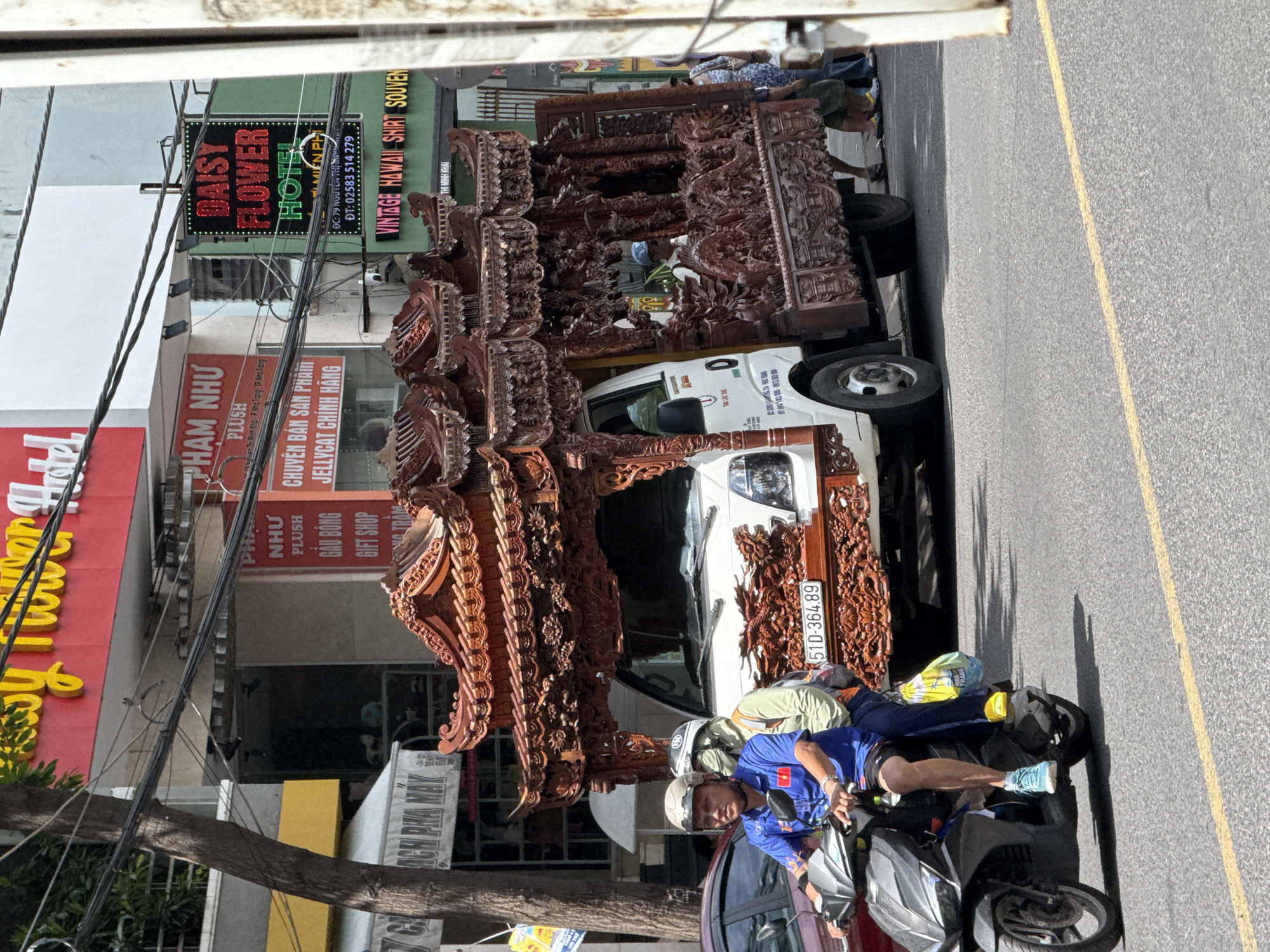 On a street in Nha Trang, Vietnam, a flatbed truck is adorned with elaborate wooden carvings, resembling a miniature temple structure. The truck carries this ornate structure, which features intricate designs, roof details, and dragon motifs. In the foreground, two individuals are riding a scooter. The driver, wearing a blue shirt and a helmet, is looking forward. The passenger behind, wearing a light green shirt, has a yellow bag at their feet. The street is paved with asphalt and marked with a yellow line. In the background, buildings and signs indicate a commercial area, with one sign for "DAISY FLOWER HOTEL" clearly visible. Power lines crisscross above the street.