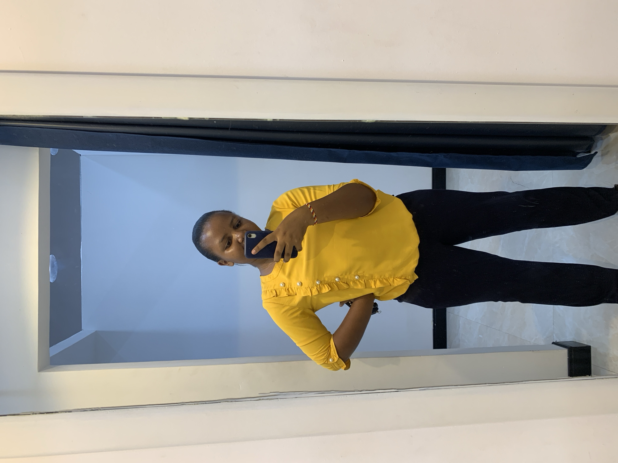 A woman is taking a selfie in what appears to be a bathroom. She is wearing a yellow shirt and black pants. The bathroom has white walls and a dark blue shower curtain rod. The location is likely in Dar es-Salaam, Tanzania, although there are no specific landmarks or indicators to confirm this.