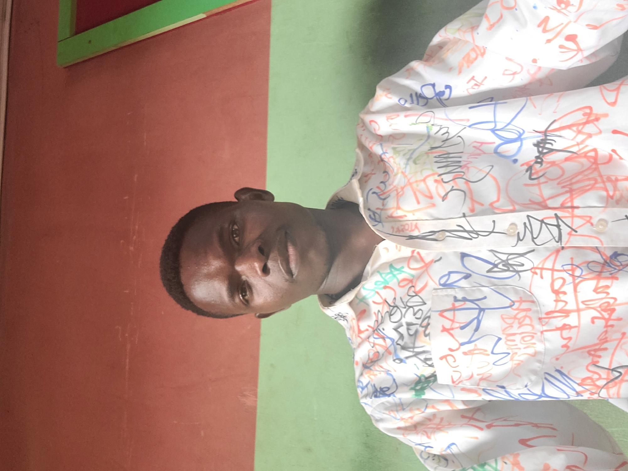 A young Black male is captured from the chest up in a portrait-style photograph. He is wearing a white, button-down shirt adorned with numerous colorful scribbles and graffiti-like markings in blue, red, orange, black, and green. A chest pocket is visible on the shirt. The man's head is turned slightly towards the camera, and he is looking directly at the viewer with a neutral expression. His skin is dark, and he has short hair.
The background is split vertically into two distinct colors: a reddish-brown on the left and a light green on the right. In the top left corner, a sliver of a structure with green and red elements is visible, possibly a window frame or part of a building. The lighting suggests it might be daytime. The overall impression is a casual, candid portrait of a young man in what appears to be an indoor or semi-outdoor setting. The image is oriented vertically, with the subject's head towards the left side of the frame.