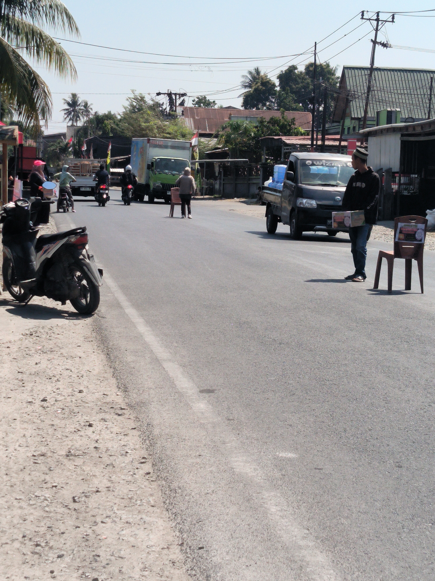 The image captures a street scene in Lemo, Indonesia, under a bright, clear sky. The road is paved, with a white line marking its center. On the left side of the road, a dirt shoulder is visible, with a black and white motorcycle parked on it. Several other motorcycles are moving on the road. A green truck with a blue and white box trailer is positioned in the middle of the road, partially obstructing traffic. To the right of the truck, a man in a black long-sleeved shirt and jeans is standing next to a dark-colored pickup truck. He is holding a box and looking towards the camera. A brown plastic chair, with a sign attached, is placed on the road next to him. In the background, buildings with corrugated metal roofs, trees, and power lines can be seen. The atmosphere appears to be that of a typical day in a busy Indonesian town.