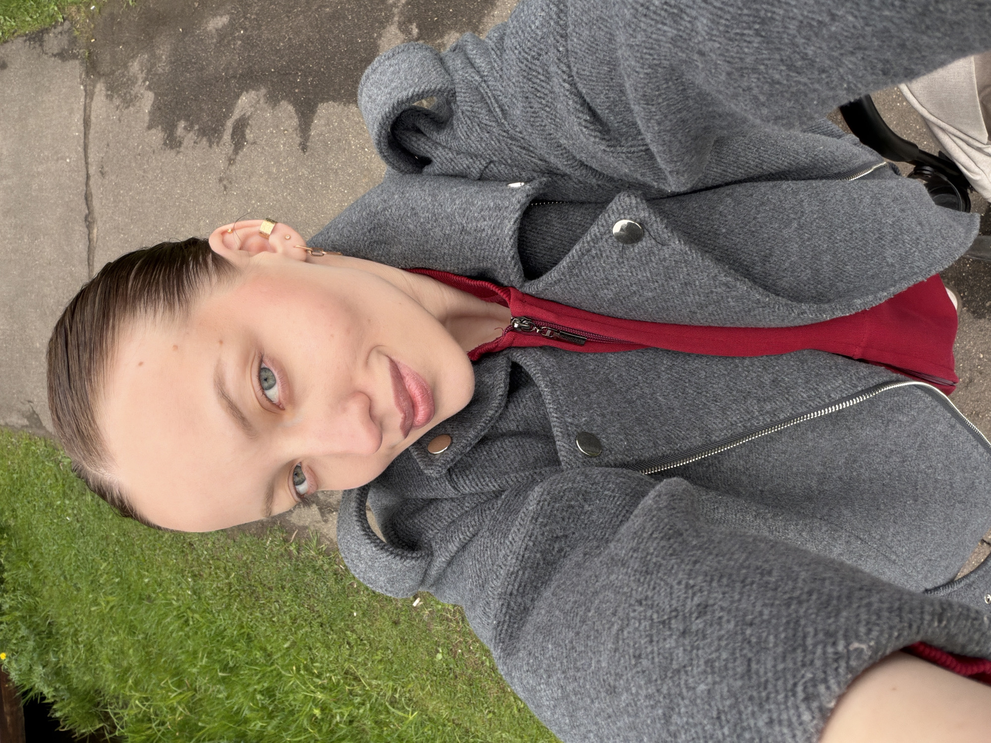 A selfie, taken from a high angle, shows a fair-skinned woman with slicked-back brown hair, light blue eyes, and light pink lipstick. She is wearing a grey woolen coat with silver buttons over a red collared shirt with a zipper. The background includes a patch of green grass and a grey concrete sidewalk. Part of a stroller is visible in the background. The photo's location is presumed to be in a similar environment as Reshetnikovo, Russia.