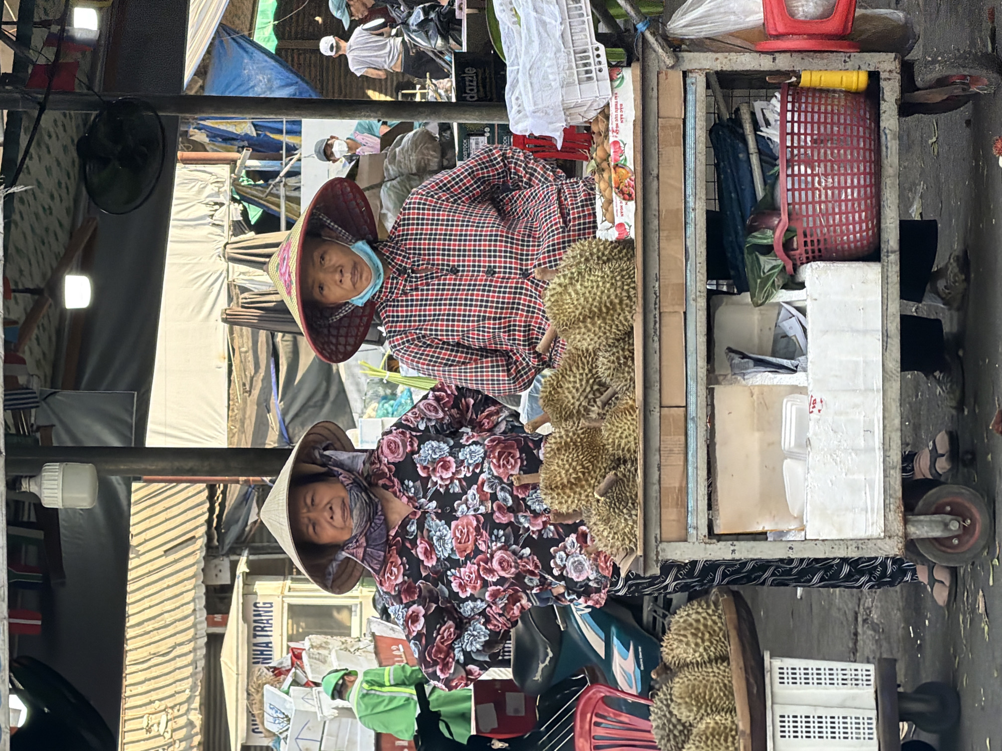 The image depicts a street market scene in Nha Trang, Vietnam. Two women, dressed in traditional conical hats and patterned shirts, stand behind a cart laden with durians. The woman on the left wears a floral shirt, while the woman on the right wears a plaid shirt. Both women have scarves or masks covering their lower faces. The cart is filled with numerous spiky durian fruits. In the background, other market stalls and vendors are visible, along with motorcycles. The environment appears to be outdoors, with makeshift roofing and structures characteristic of a busy Asian market. The overall impression is one of commerce and daily life in a vibrant urban setting.